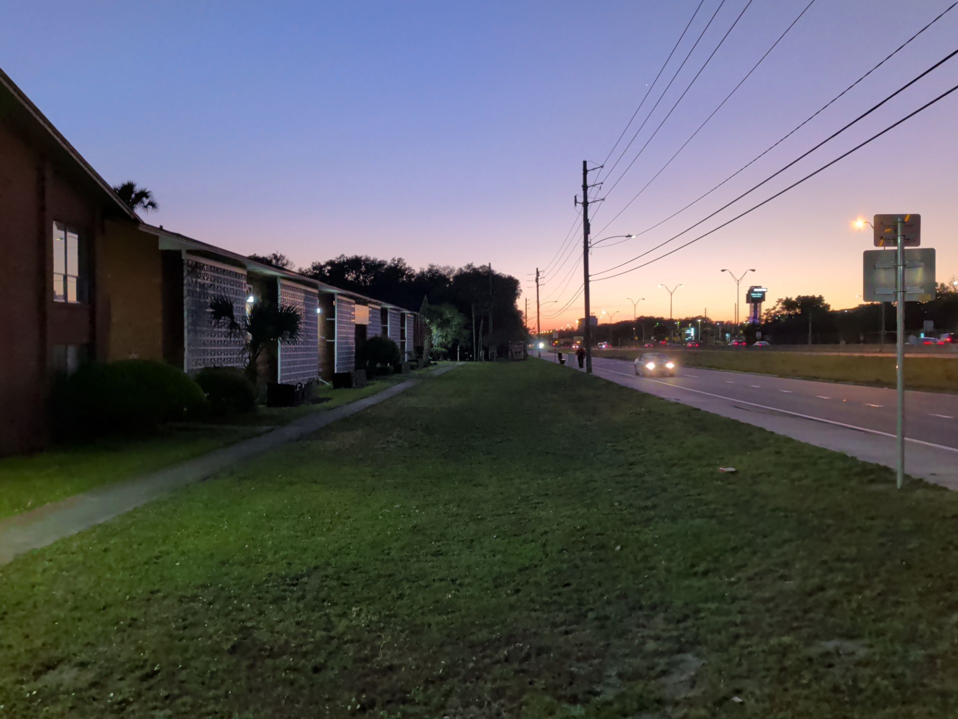 Grassy strip beside apartment buildings and a road at sunset, with cars and utility poles along the street.