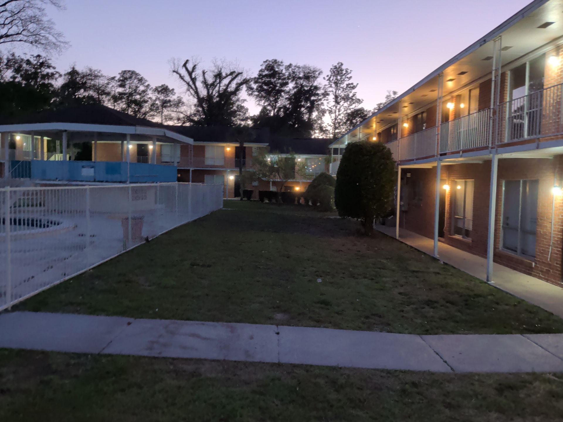 Apartment courtyard at dusk with lit balconies, white fence, and grassy central lawn