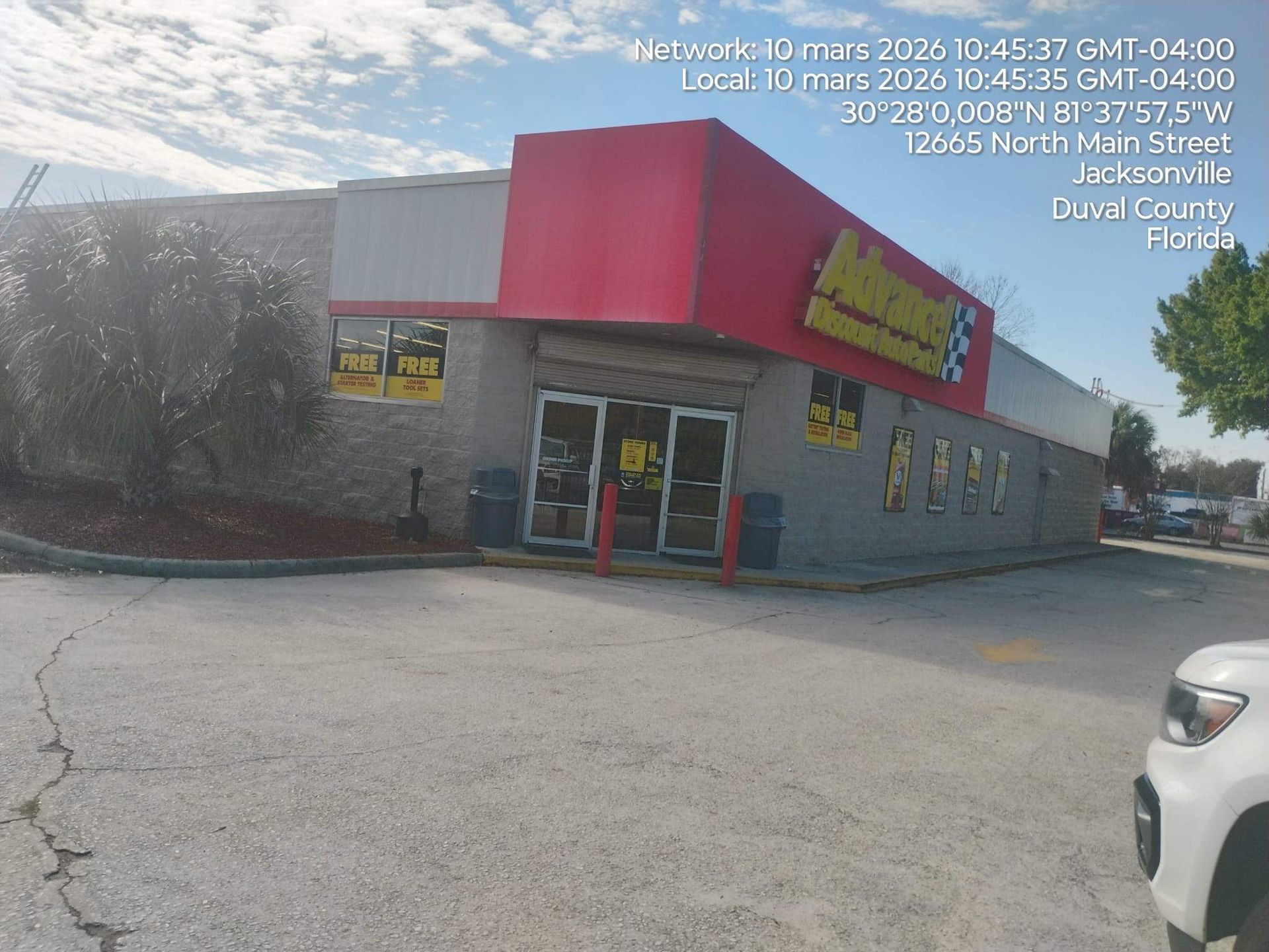 Street-view of a gray-and-red convenience store with a yellow sign and parking lot, partially obscured by trees