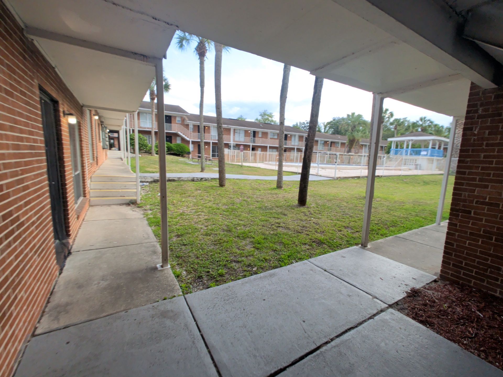 Covered walkway beside a brick building, overlooking a grassy courtyard and apartment buildings beyond.