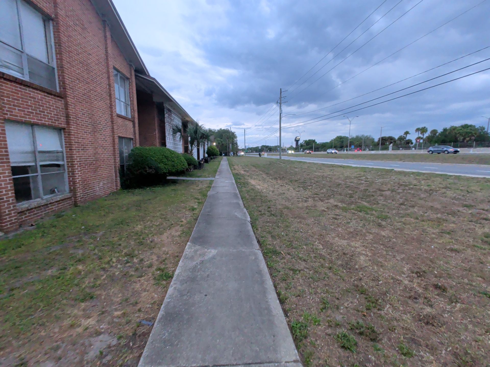 Sidewalk beside a brick building and grassy roadside under a cloudy sky