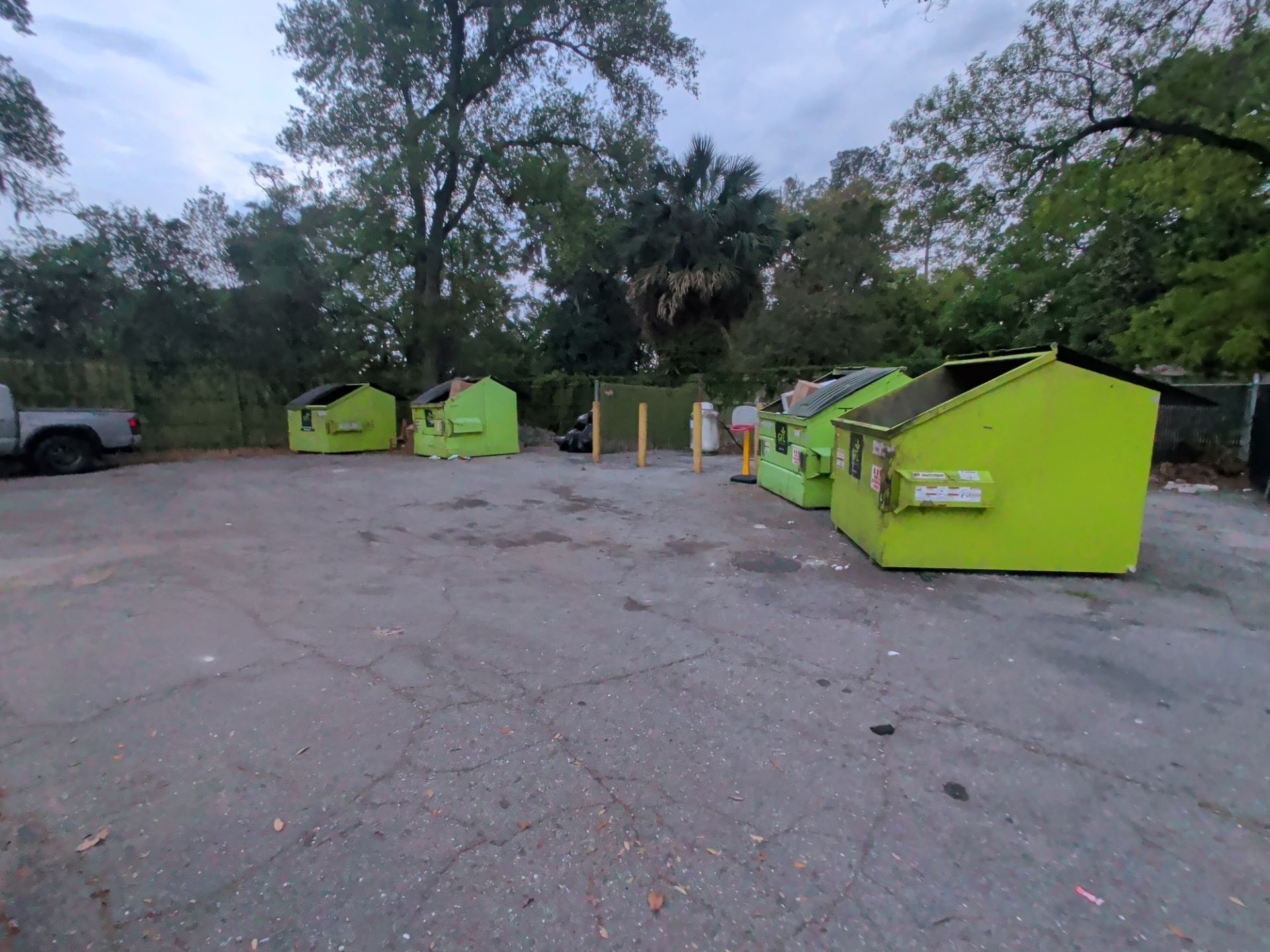 Bright green dumpsters and a garbage truck in a wooded parking lot at dusk