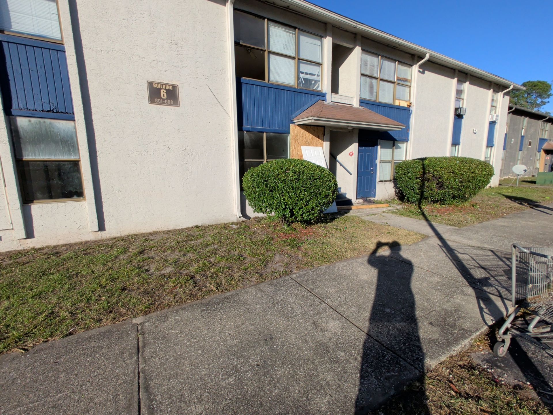 Apartment building exterior with blue awnings, shrubs, and a sidewalk in sunlight