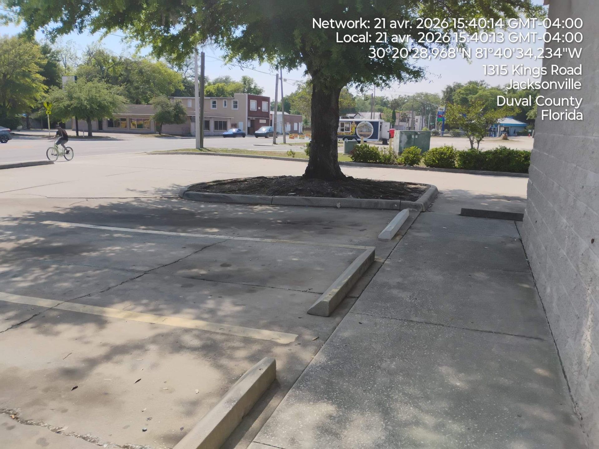 Empty sidewalk and parking area beside a building, with a tree and low curbs in bright sunlight.