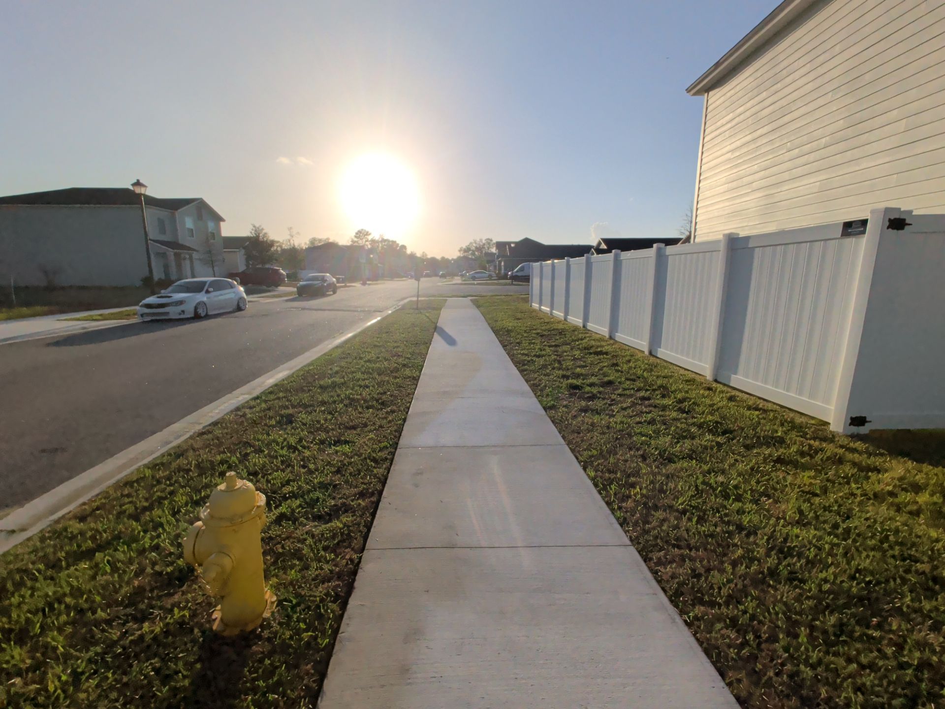 Sunlit suburban sidewalk beside a street, white fence, parked car, and yellow fire hydrant at dusk