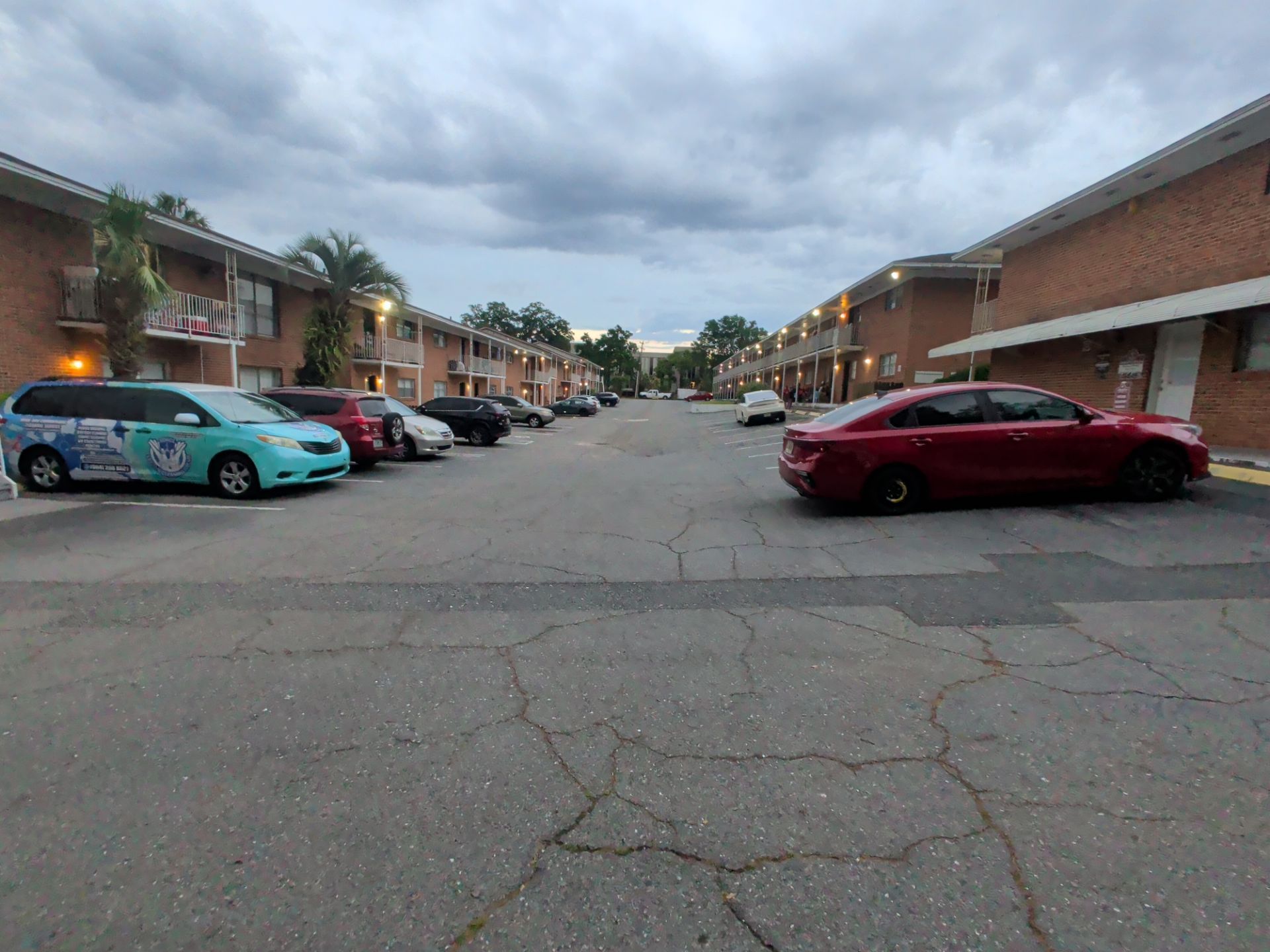 Apartment parking lot with cars along both sides under a cloudy sky, including a turquoise car and red sedan.