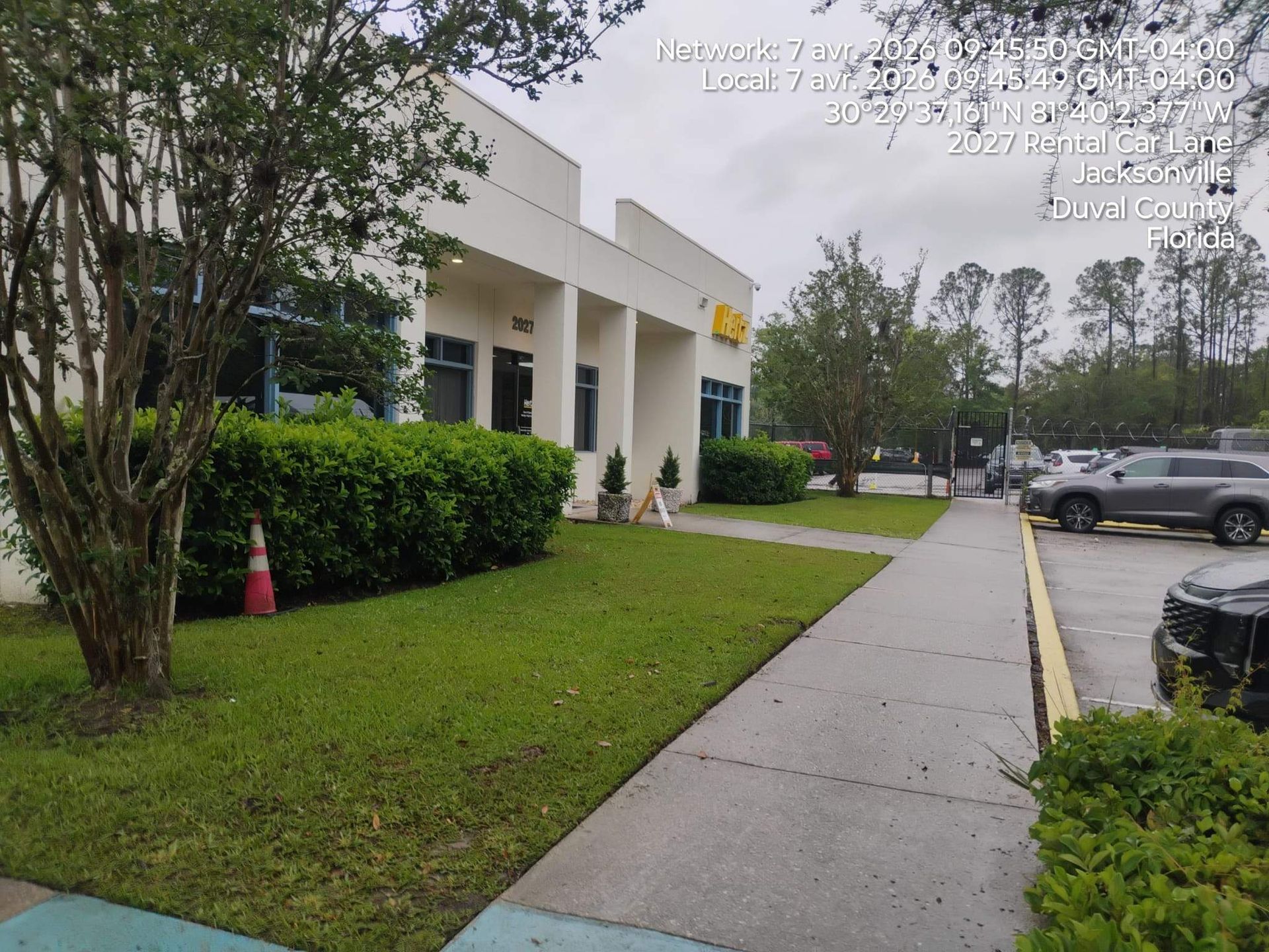 Sidewalk beside a white office building, with hedges, trees, parked cars, and a cone on the lawn.