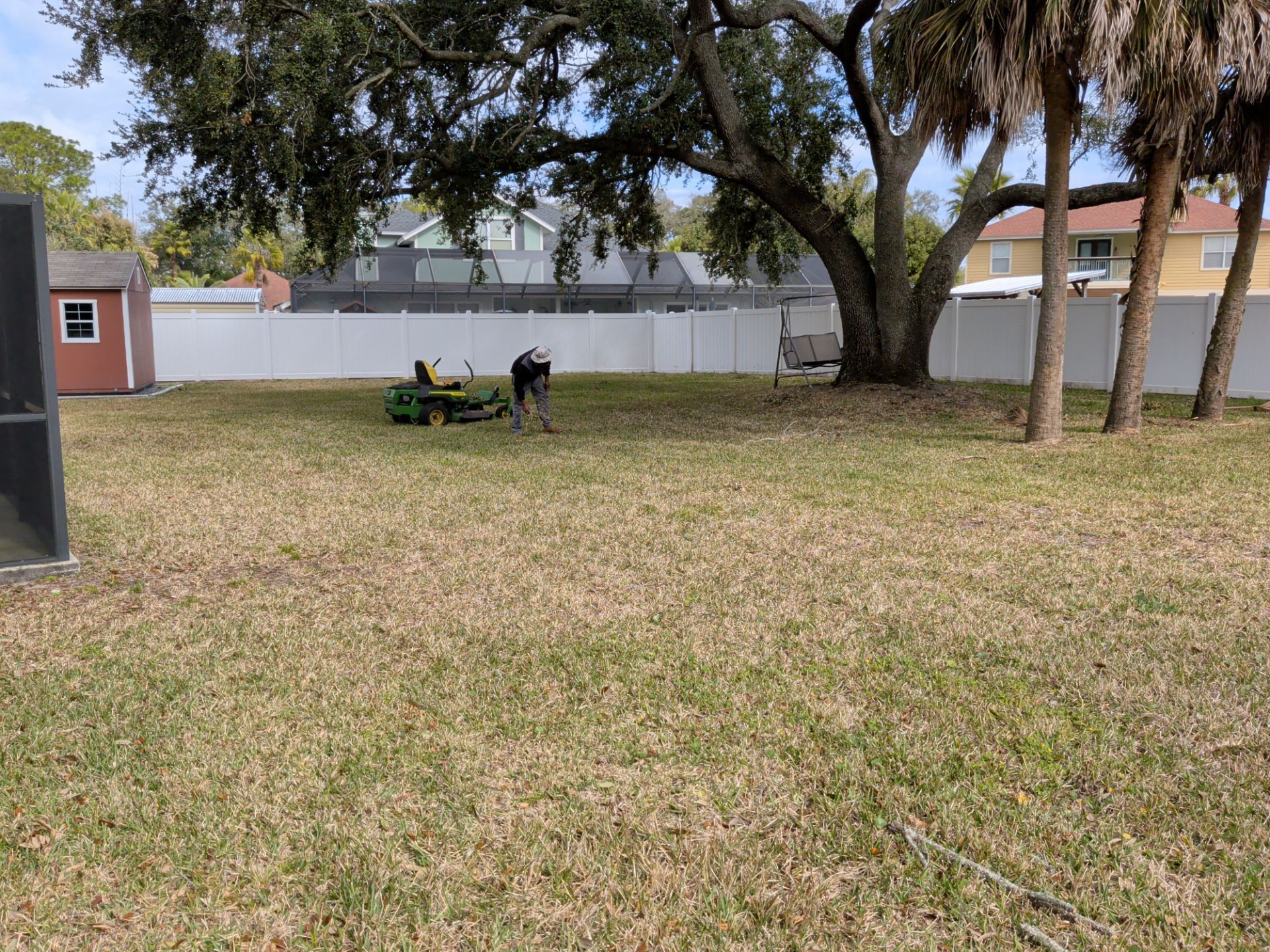 Two people sitting on a bench in a grassy yard under large trees, with a white fence in the background.