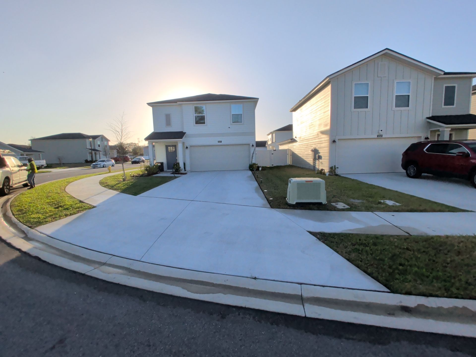 Suburban street with two houses, driveway, sidewalk, and parked cars at sunset.