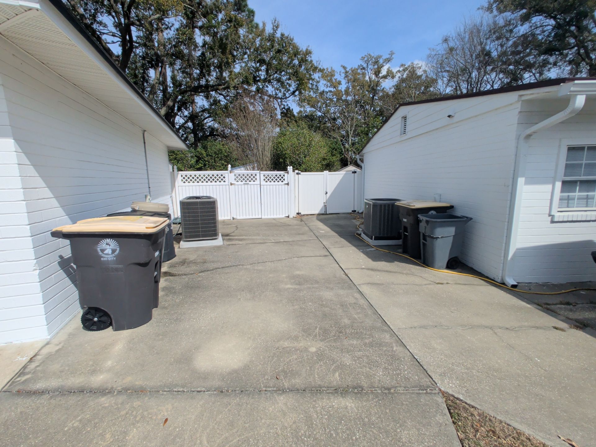 Concrete driveway between white buildings with trash bins and a closed white gate.