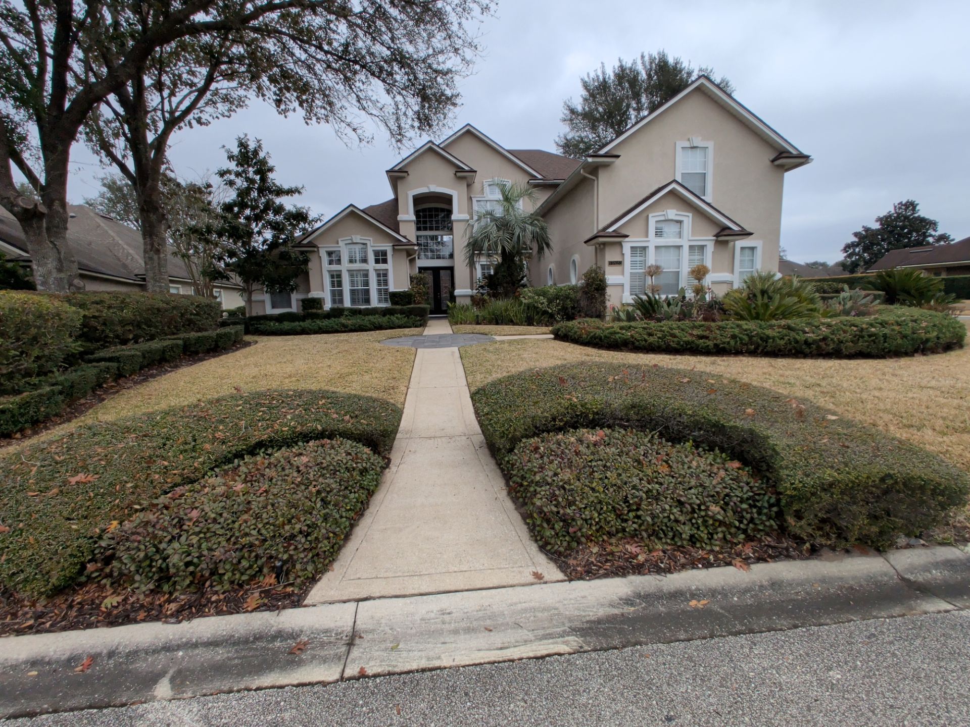 Suburban house with beige exterior, manicured hedges, and a concrete walkway leading from the street.