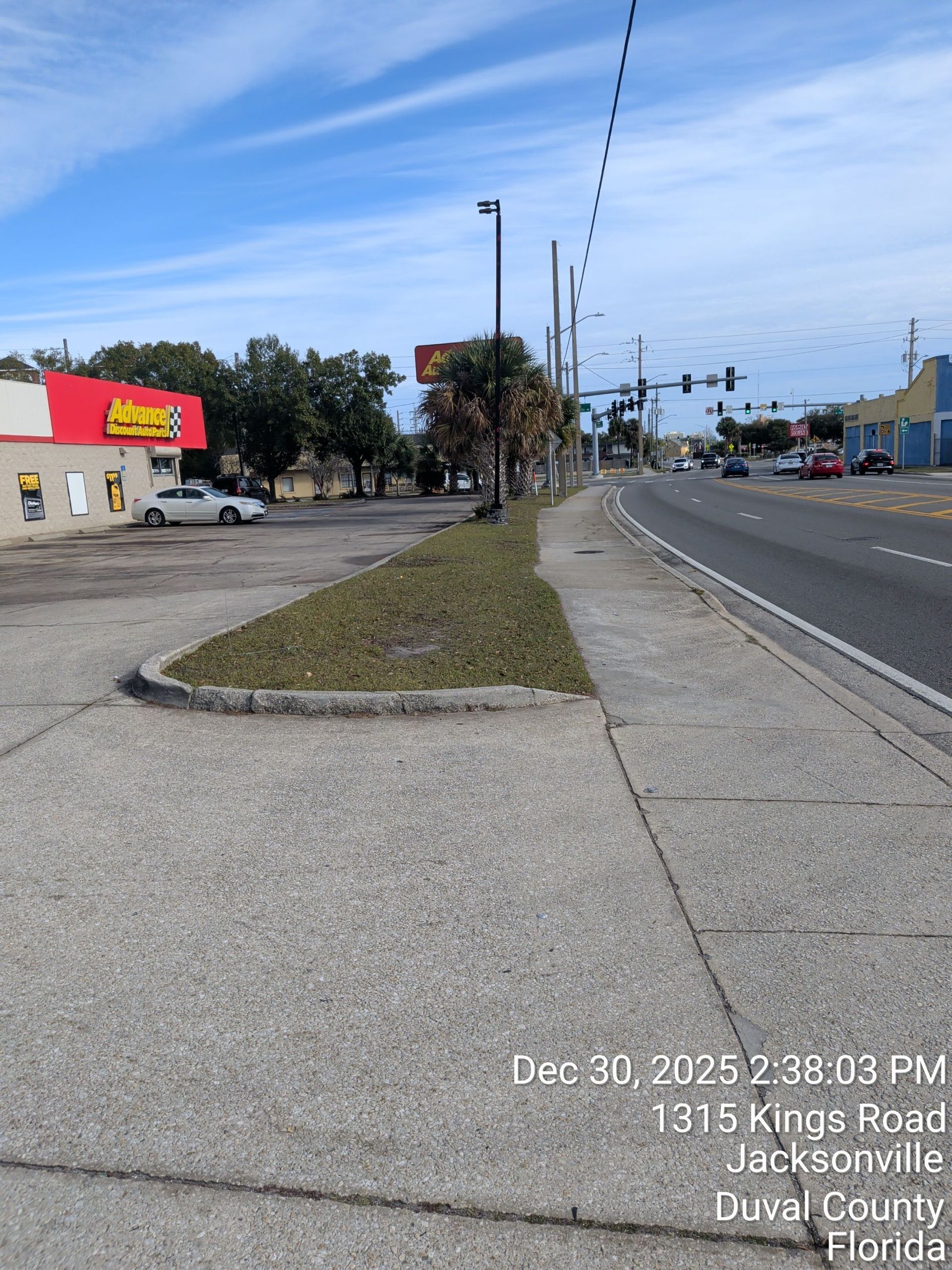 Wide roadside sidewalk beside a busy street, with trees, buildings, and a grassy median under a clear sky.