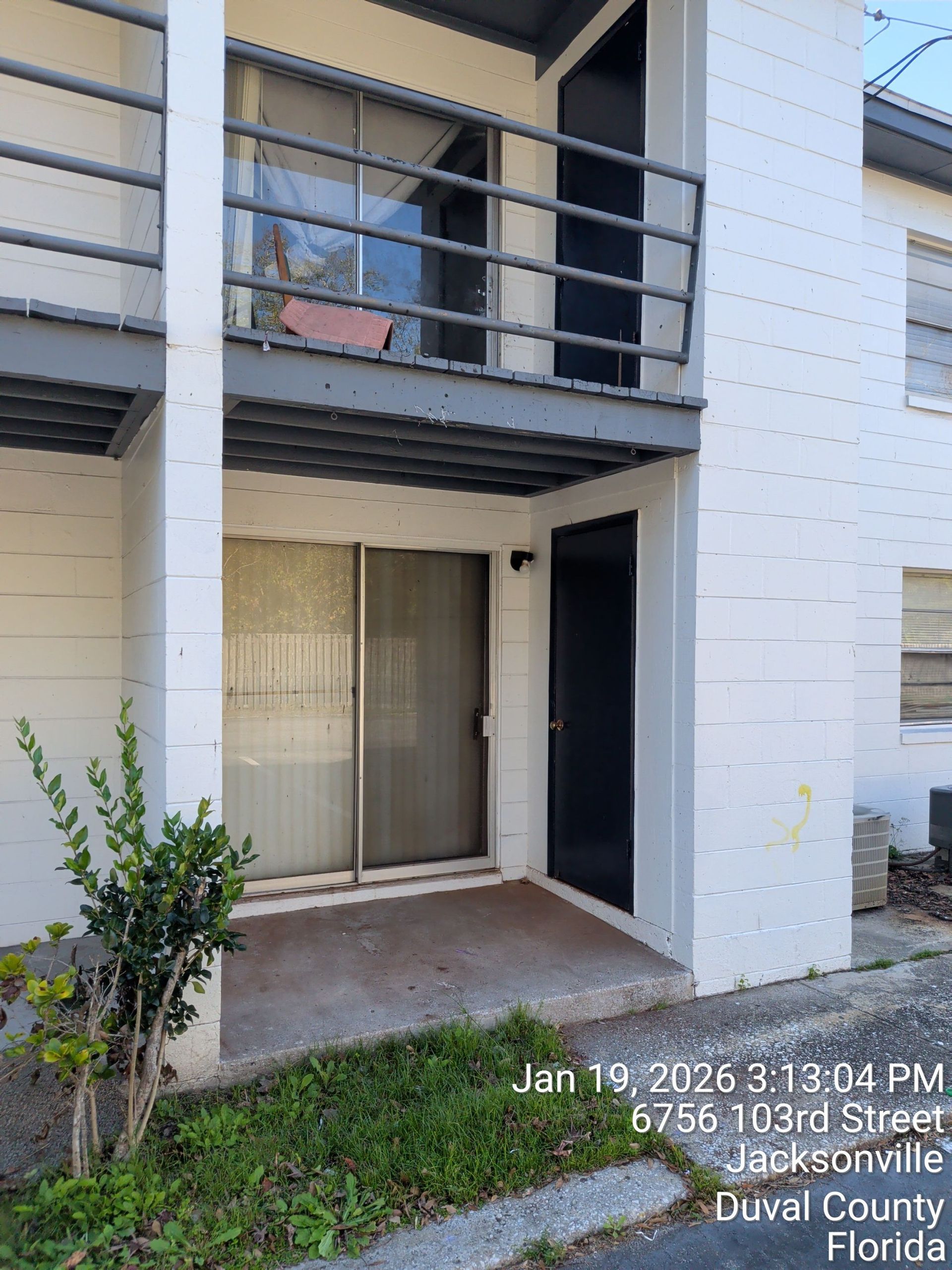Apartment patio with sliding glass door, balcony above, and small shrub beside a walkway.