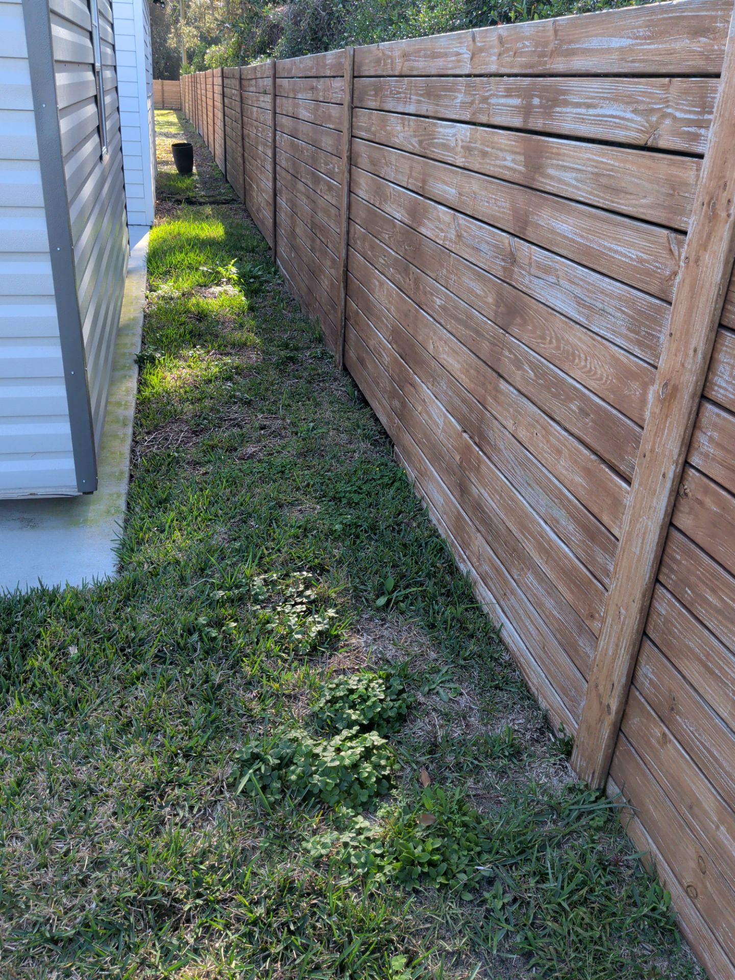 Narrow grassy side yard beside a white house and wooden fence, with a person in the distance.