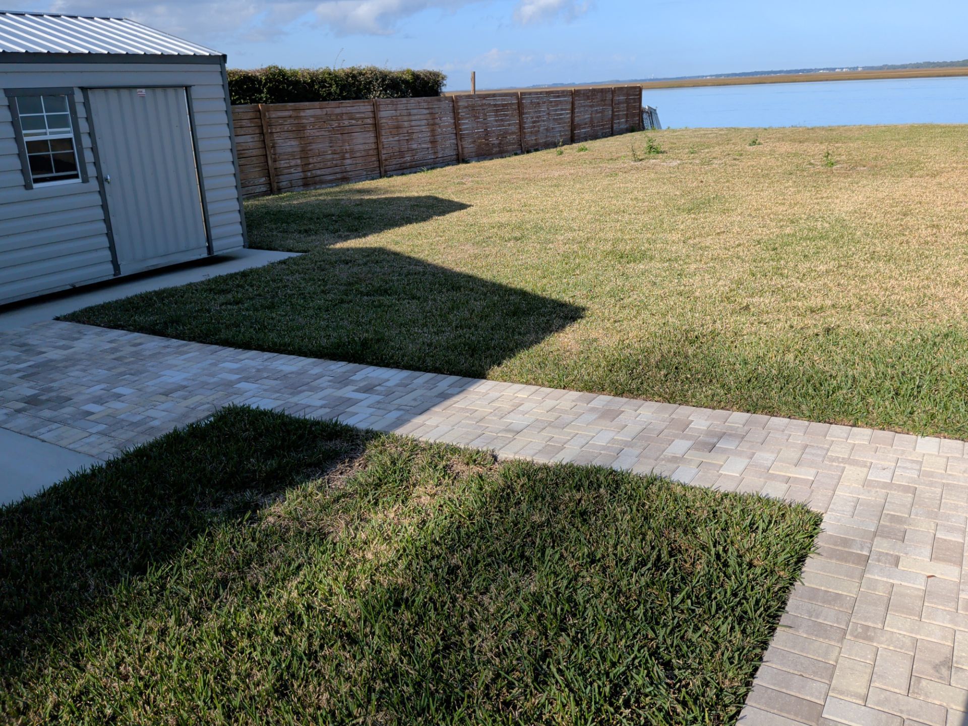 Backyard with green lawn, stone patio, and small shed beside a brick fence under a clear sky