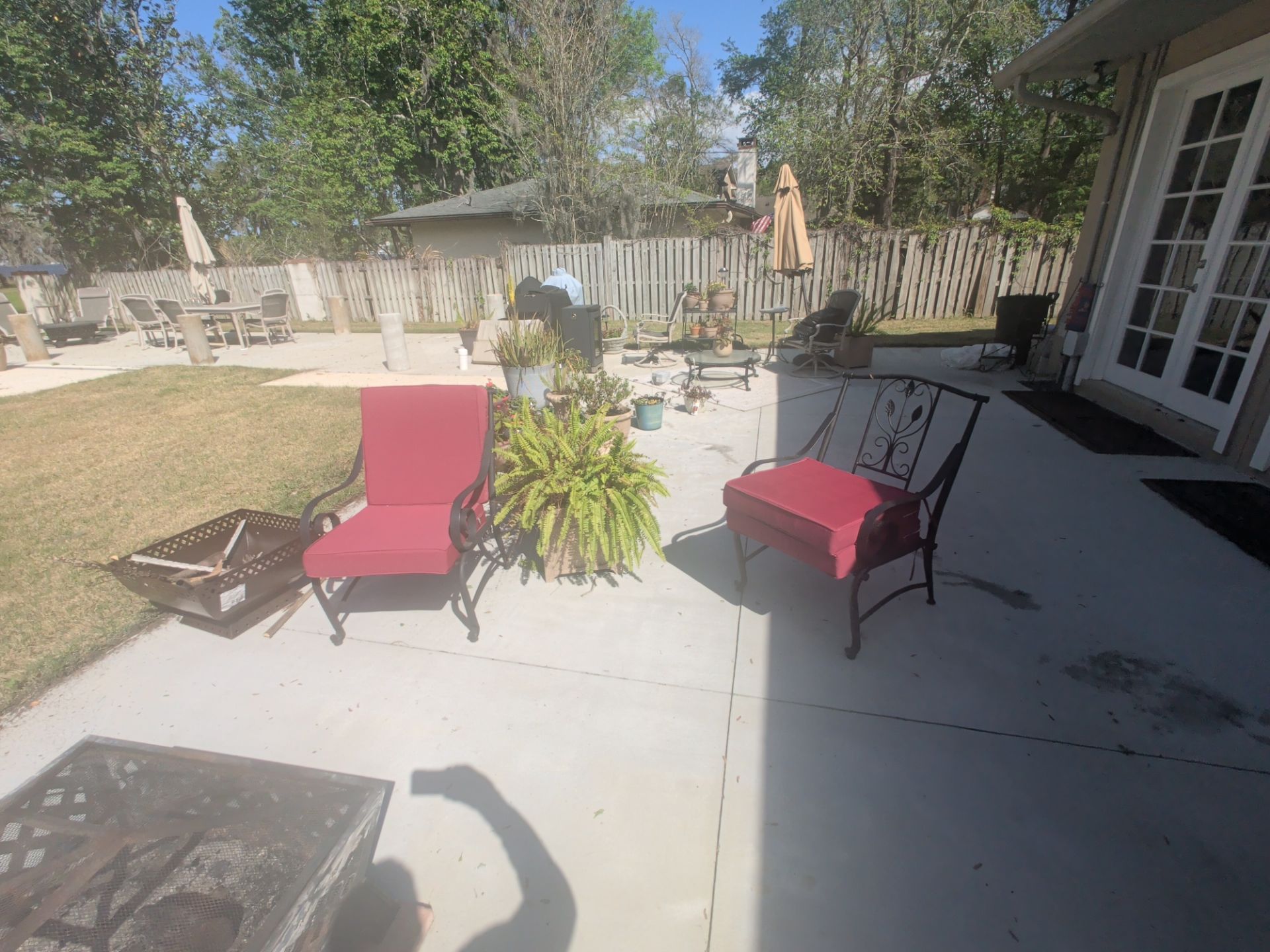 Patio with red chairs, potted plant, and fire pit beside a house and fenced backyard in sunlight