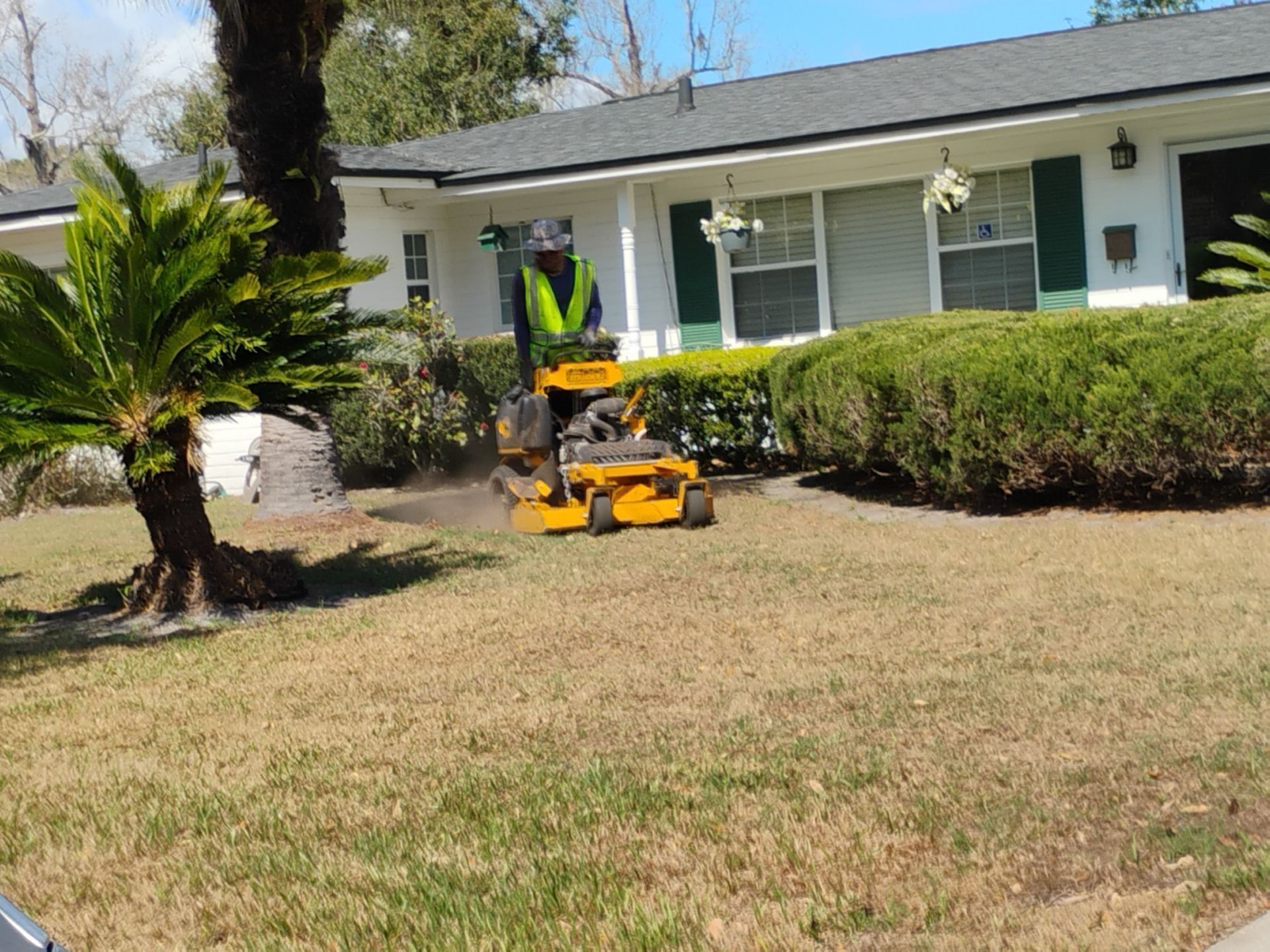 Person mowing a dry front lawn with a yellow riding mower in front of a white house