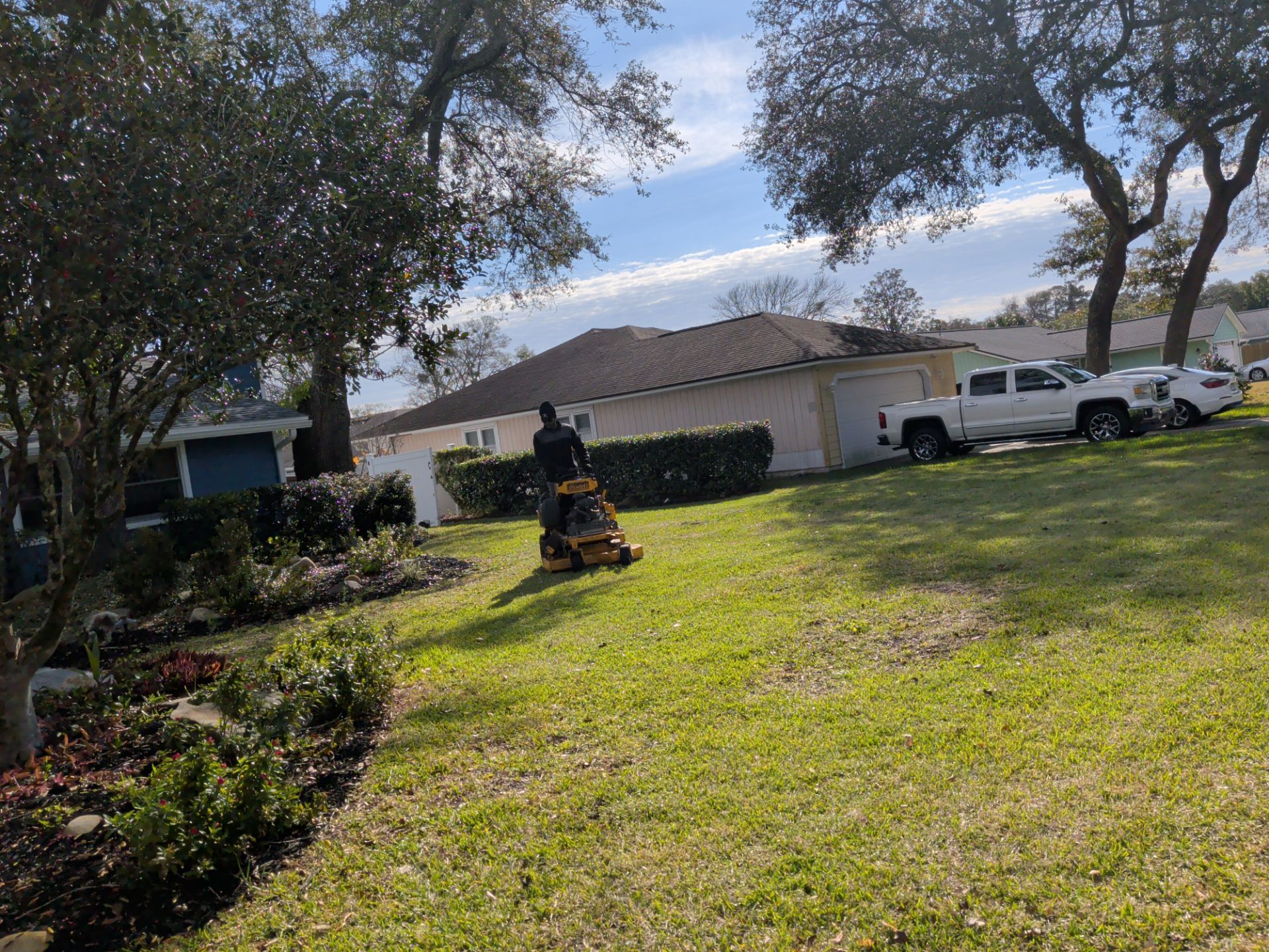 Person mowing a grassy lawn in front of a suburban house with trees and parked vehicles