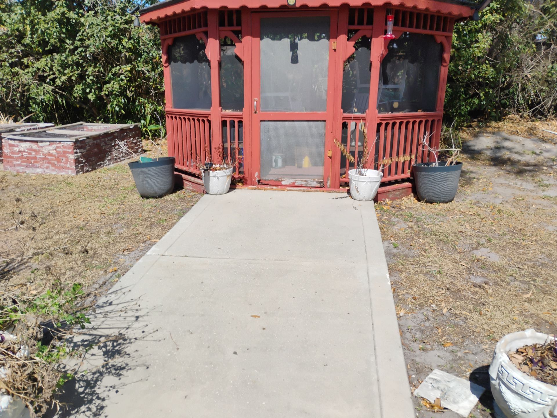 Red gazebo at the end of a concrete walkway in a dry yard, with potted plants on either side.