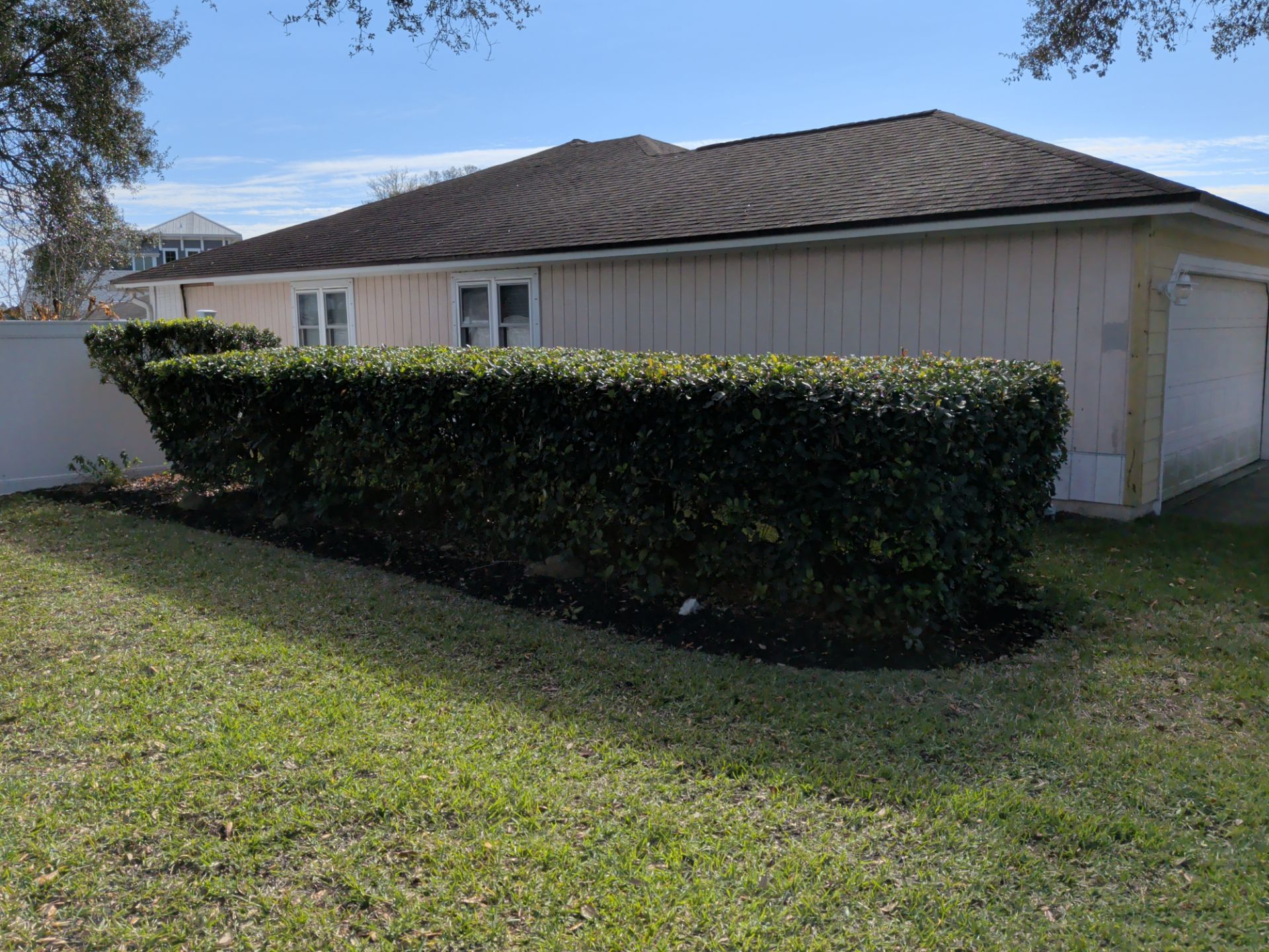 Single-story beige house with a dark roof, hedge, and grassy yard under a blue sky