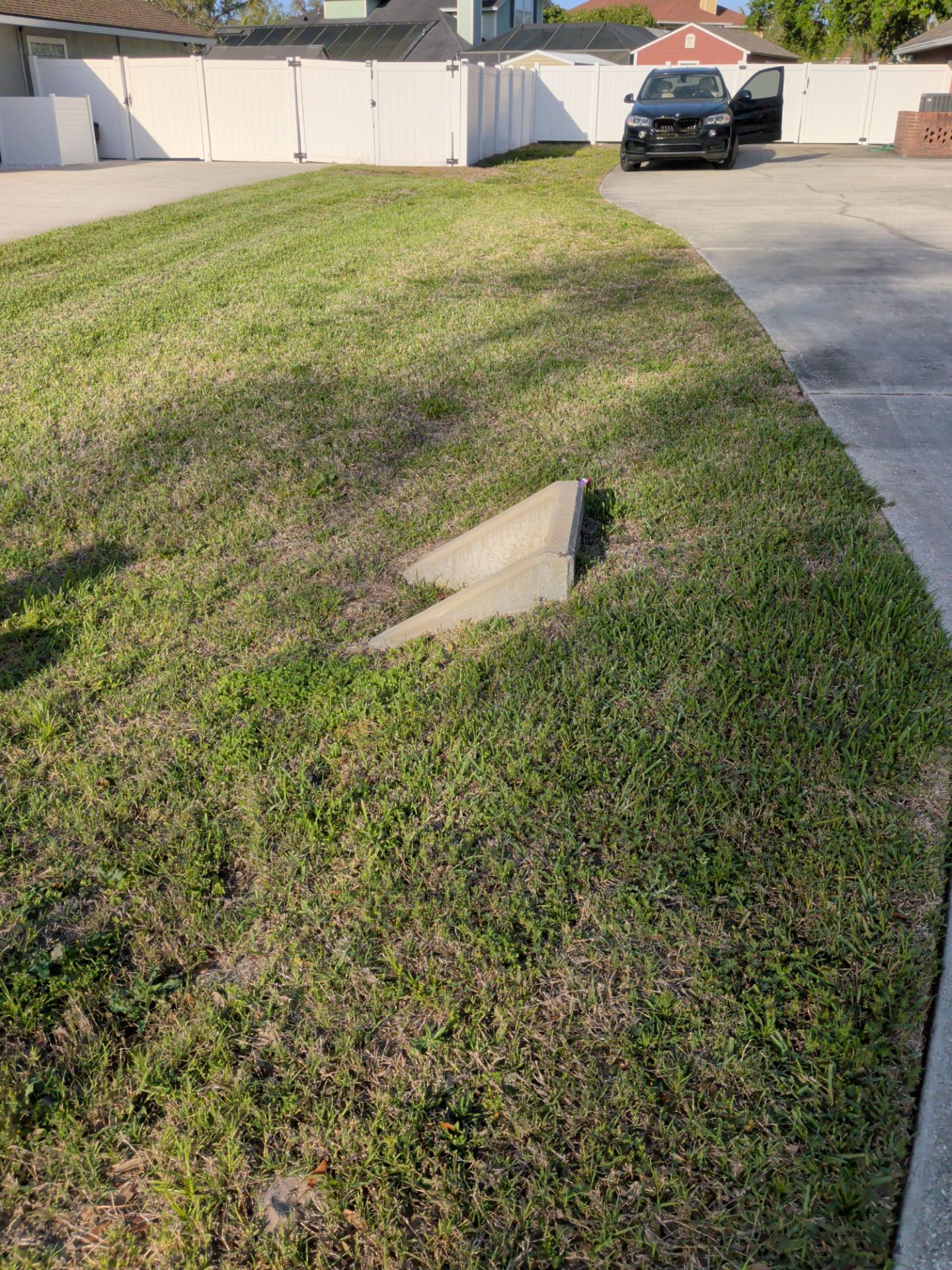 Grass-covered roadside curb with a concrete storm drain opening beside a driveway and parked car.