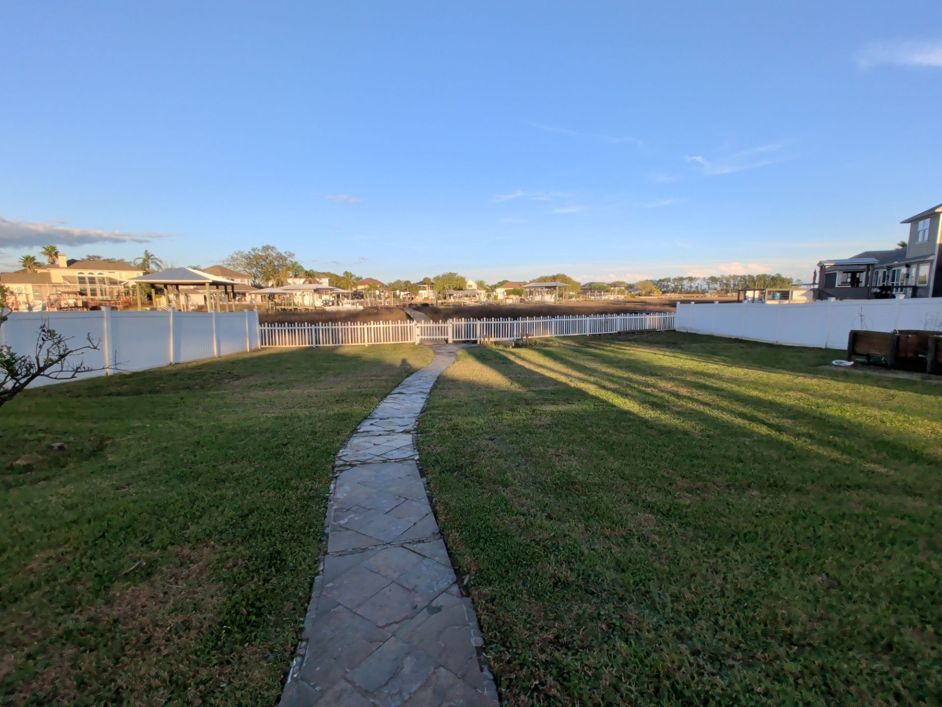 Curved stone path through a grassy waterfront lawn at sunset, with houses and a blue sky in the distance