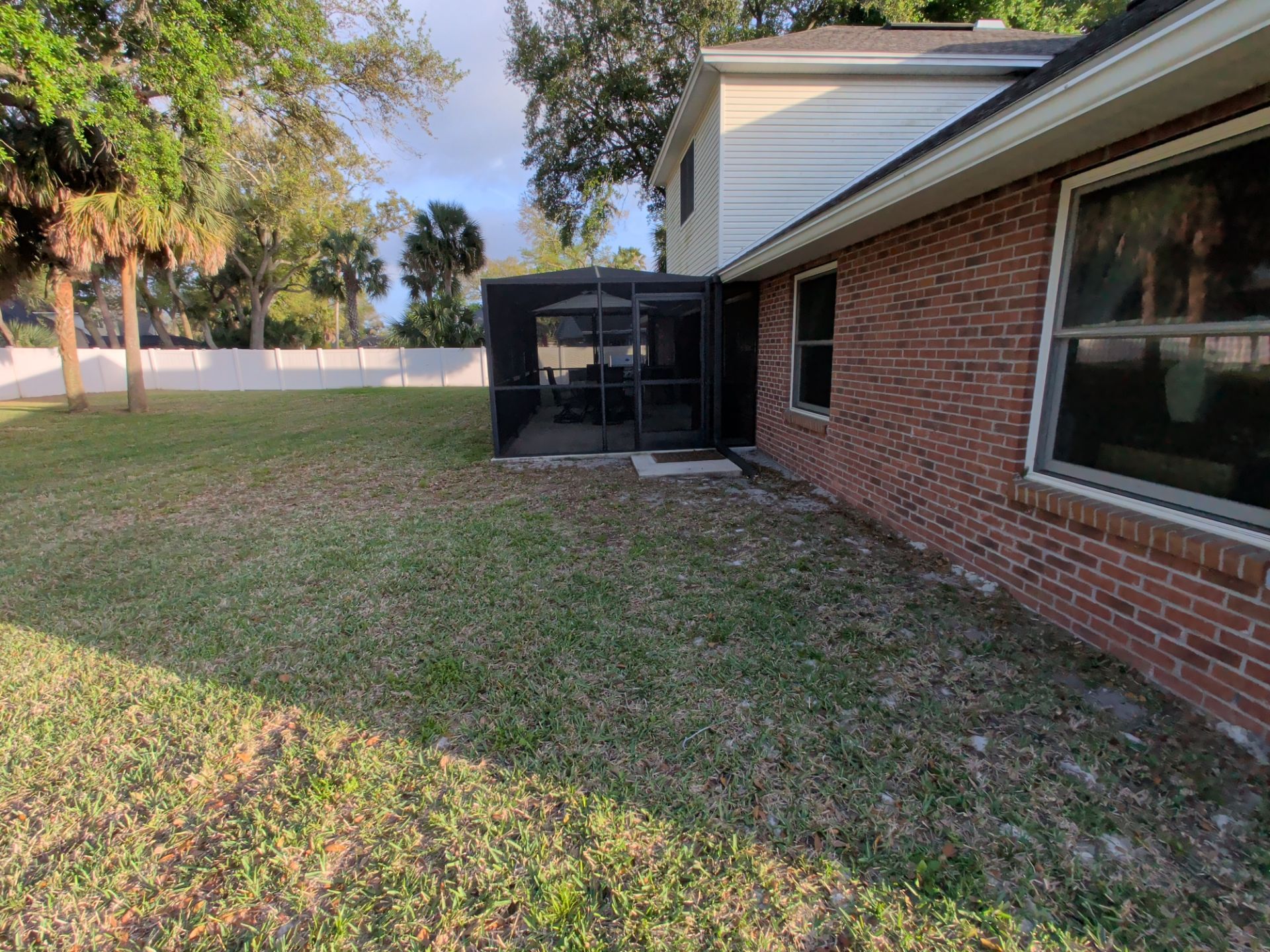 Backyard beside a brick house with screened patio, grassy lawn, and trees in the background