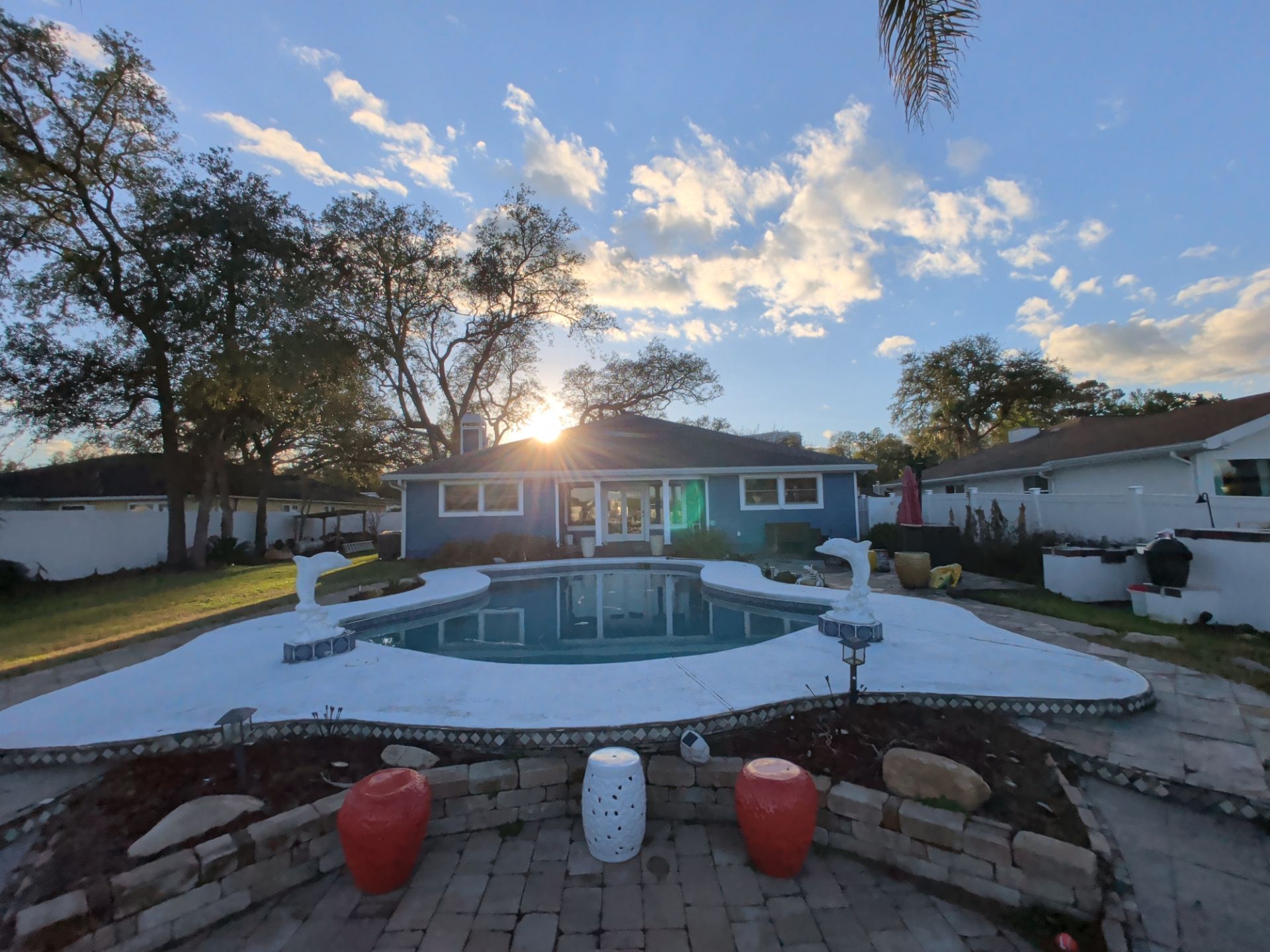 Backyard pool with white deck and red pots at sunset, house centered behind it.