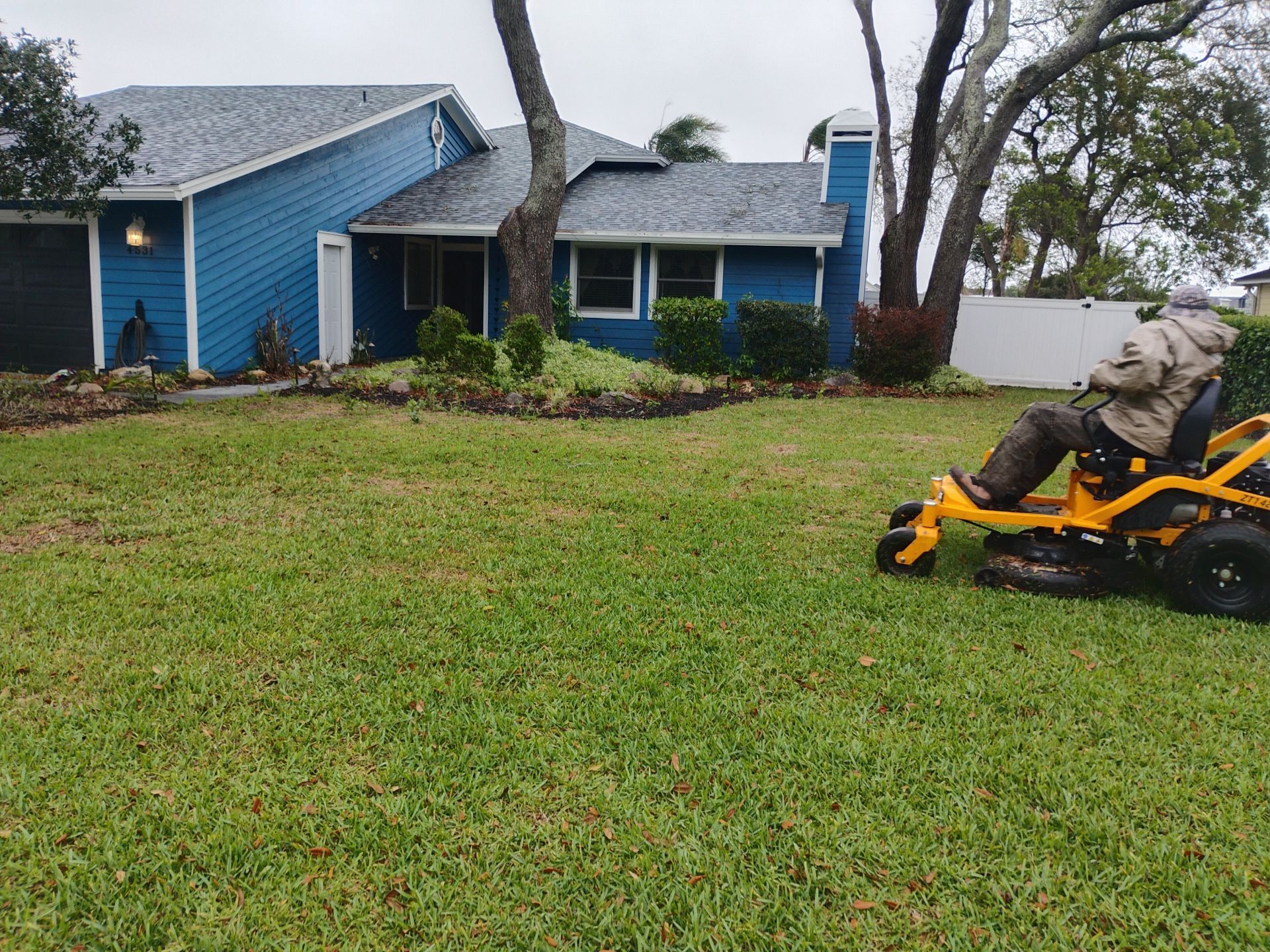 Yellow lawn mower on a grassy yard in front of a blue house with trees and a white fence.