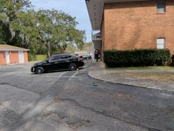 Black car parked near a brick building. Trees and a small shed are in the background.