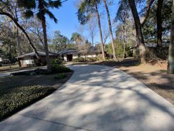 Long concrete driveway leads to a single-story home with surrounding trees and a clear blue sky.
