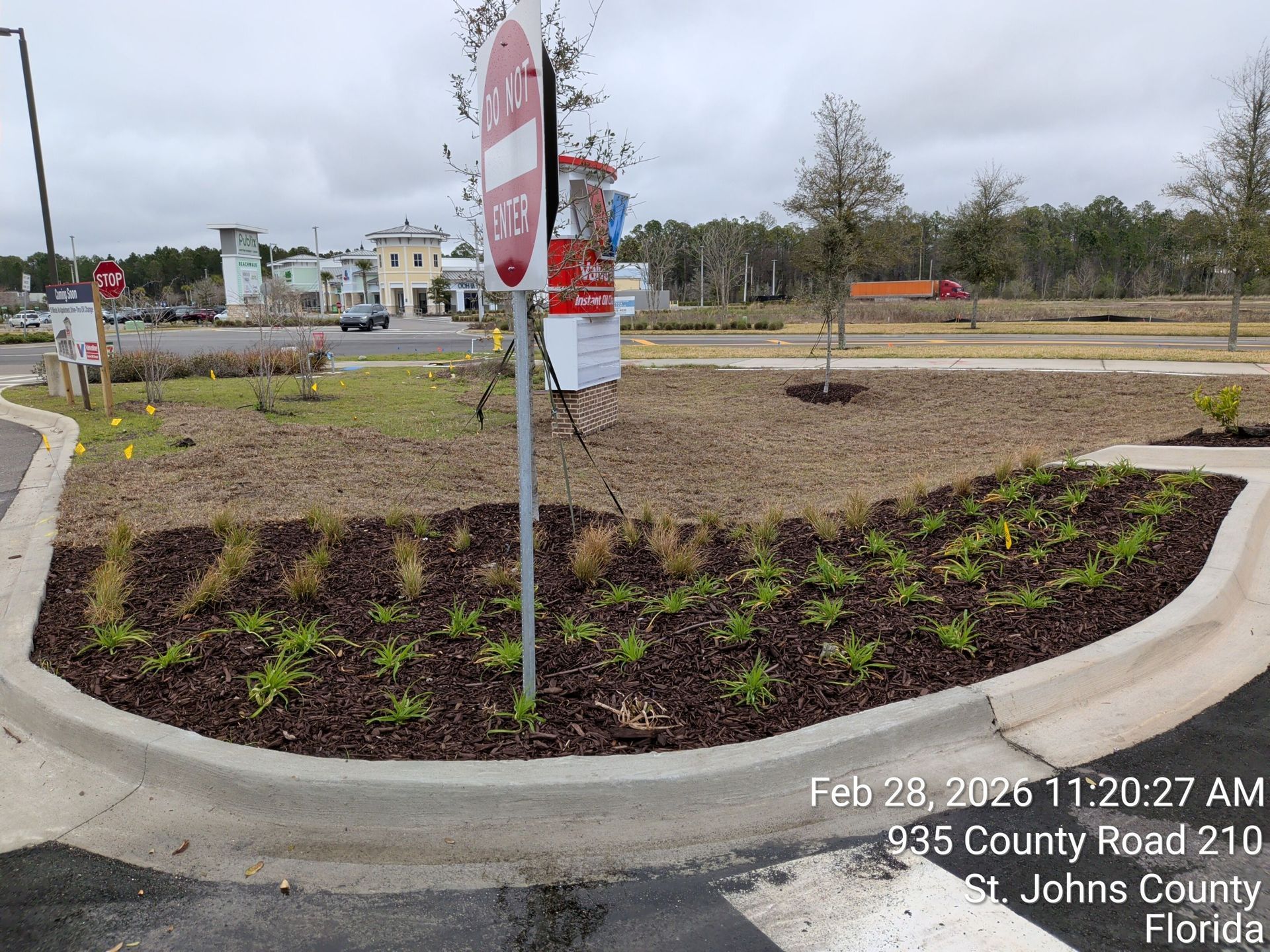 Curved flower bed with small green plants around a metal signpost on a cloudy roadside