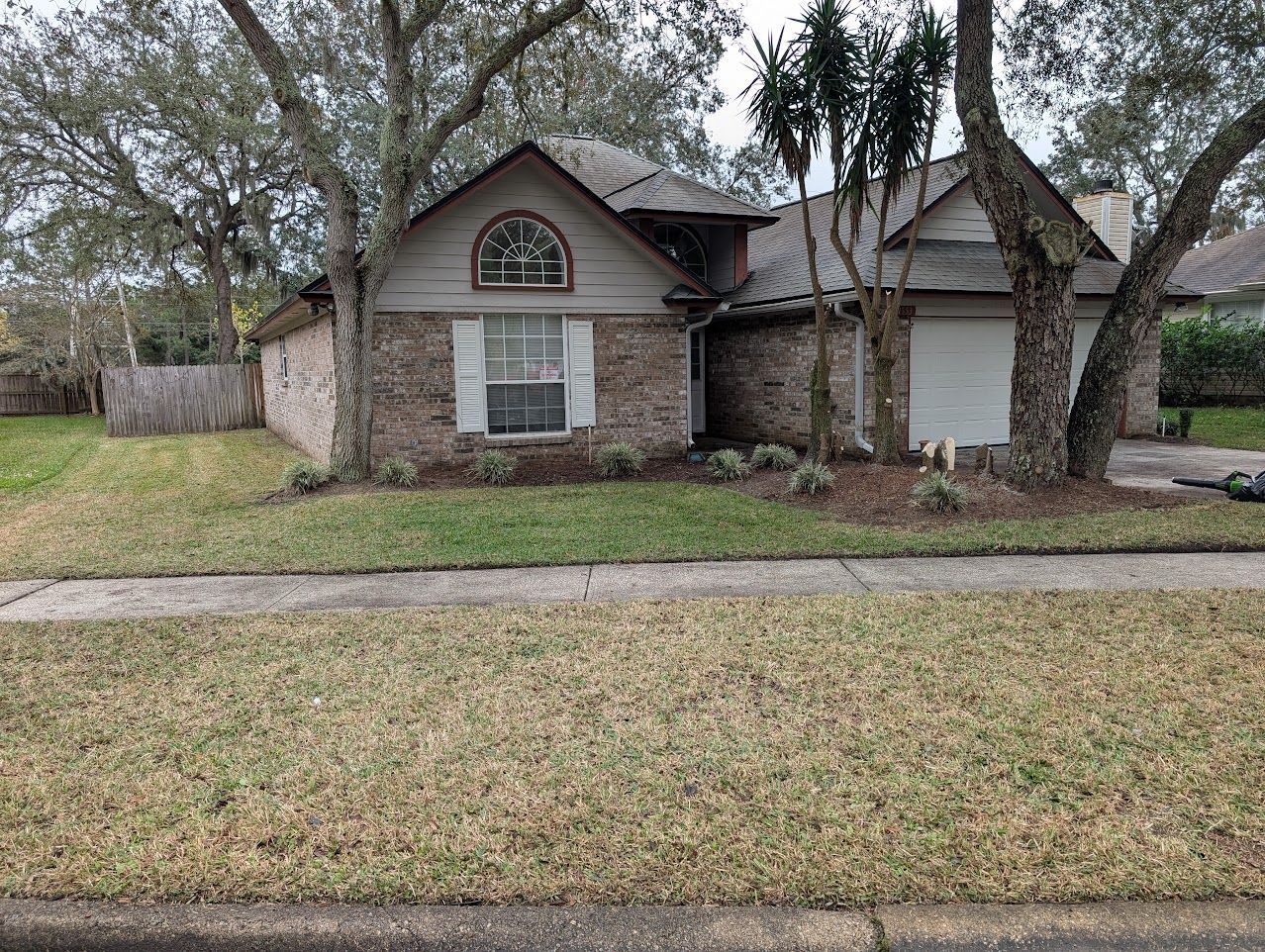 Suburban house with brick facade, arched window, and attached garage. Brown roof, green lawn, trees.
