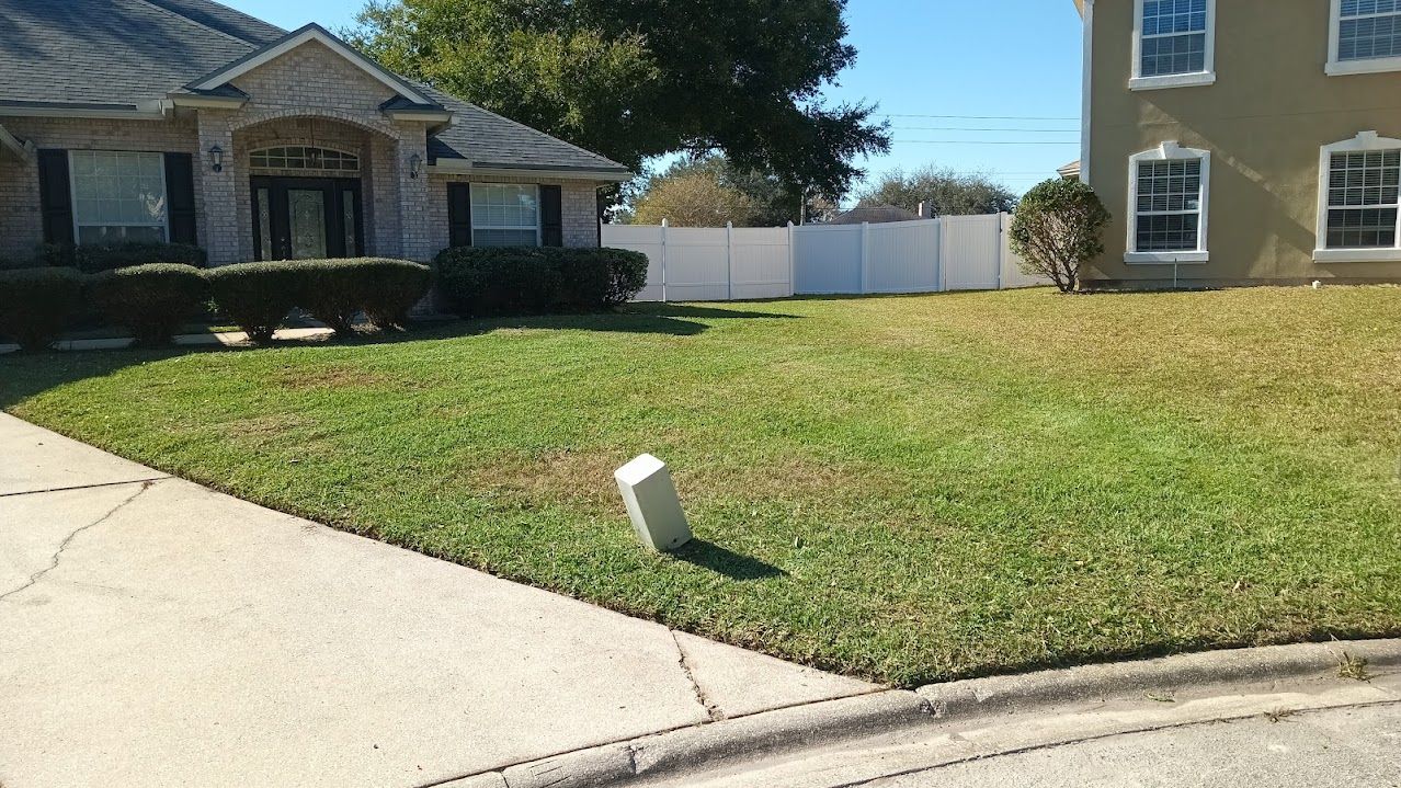 A house with a front lawn, sidewalk, and a white fence in the background. The grass is partly green and brown.