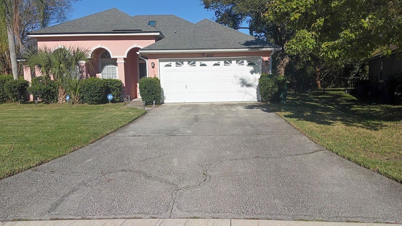 Pink house with gray roof, white garage door, and a textured concrete driveway on a sunny day.