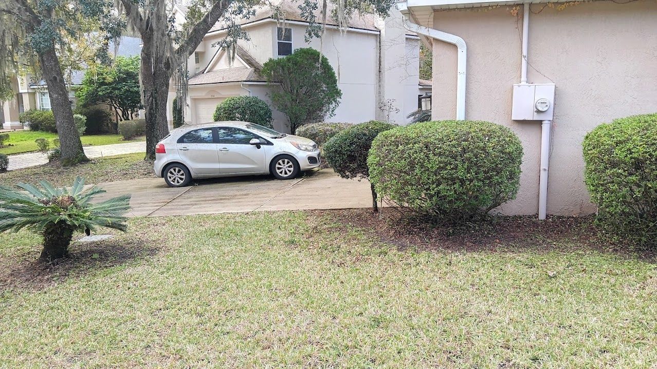 A white car parked on a driveway in front of a house, with green bushes and grass in the yard.