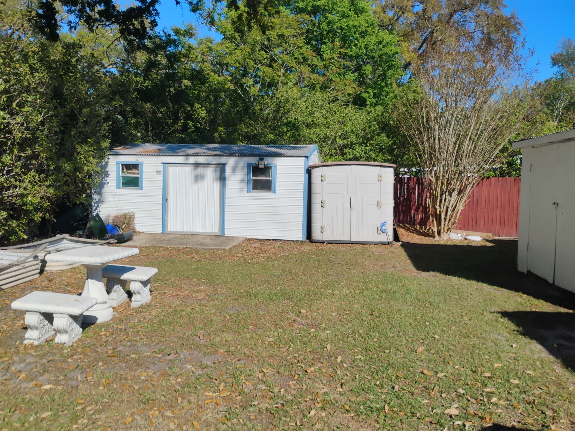 Backyard with two white storage sheds, a picnic table, and trees in the background.