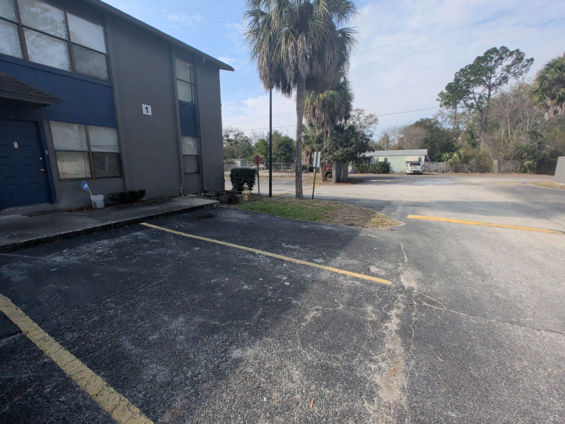 Apartment parking lot with blue-gray building, empty spaces, palm tree, and yellow curb markings
