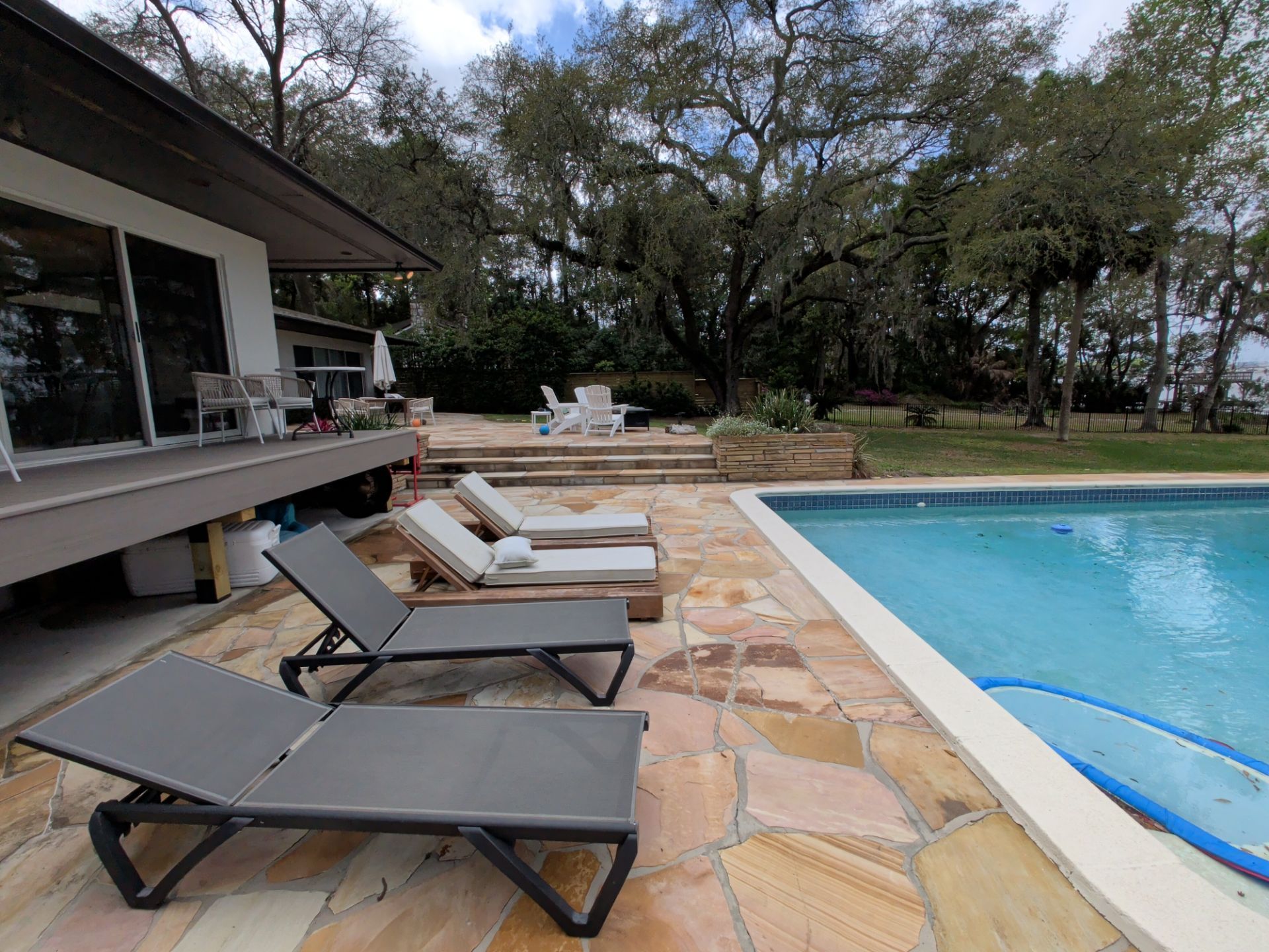 Poolside patio with lounge chairs beside a blue swimming pool and a house deck under trees