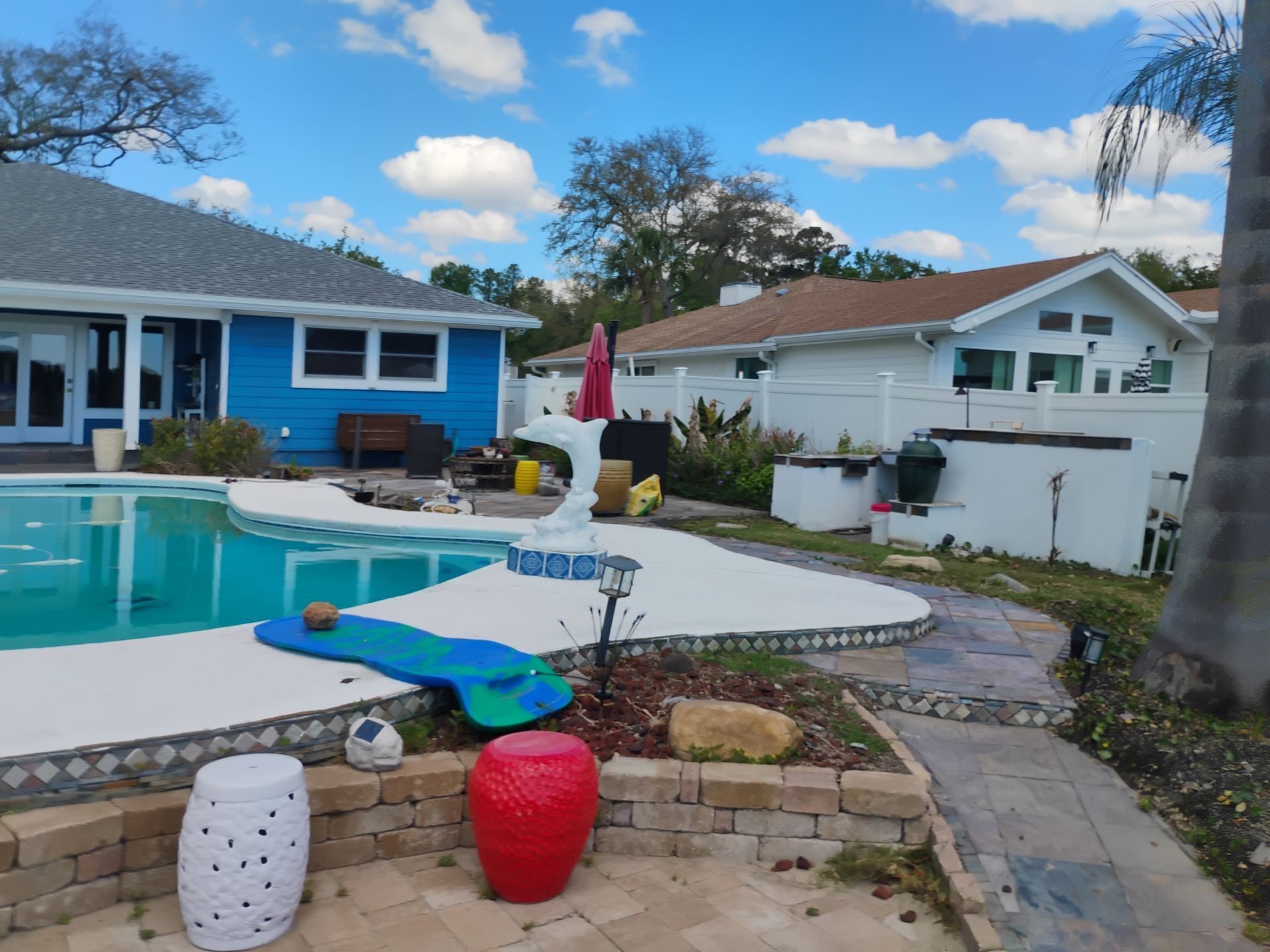 Backyard pool with blue house, white fence, patio chairs, and red and white stools by the water