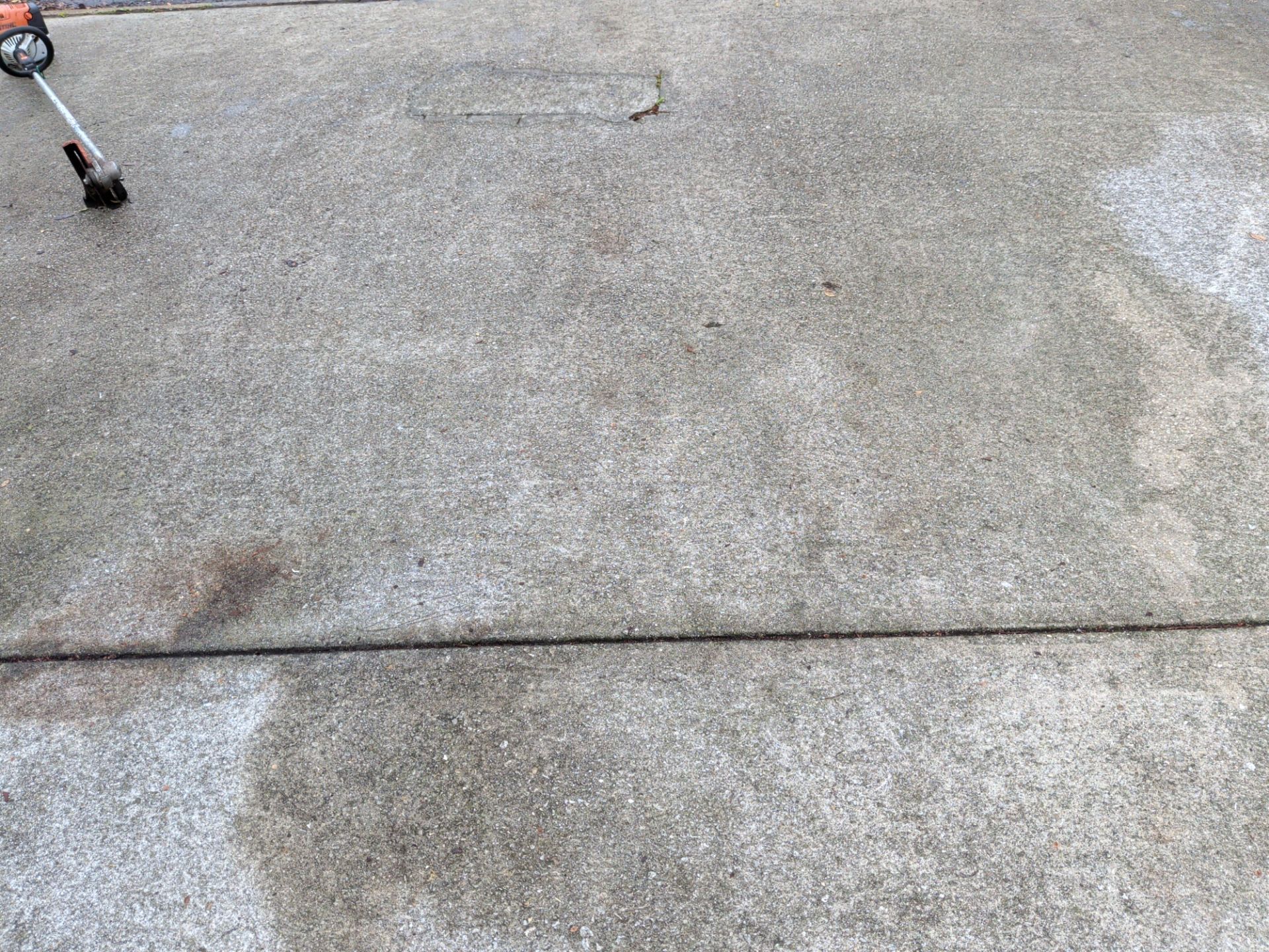 A person is using a weed whacker on a concrete surface, with the tool visible in the upper-left corner.