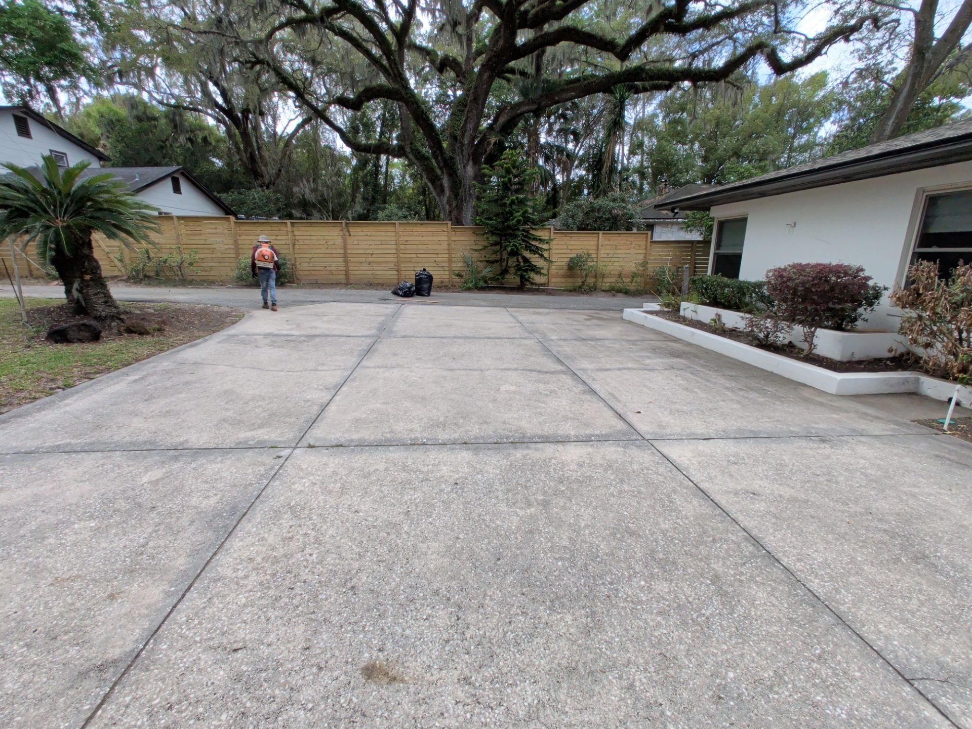 Wide concrete driveway with a person standing near a wooden fence and a house on the right.