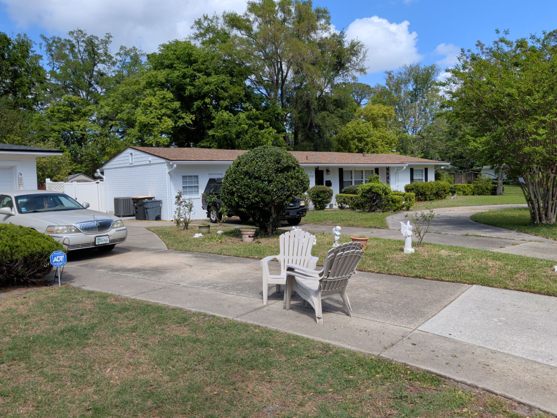 Suburban house with driveway, parked car, lawn chairs, and trees under a partly cloudy sky