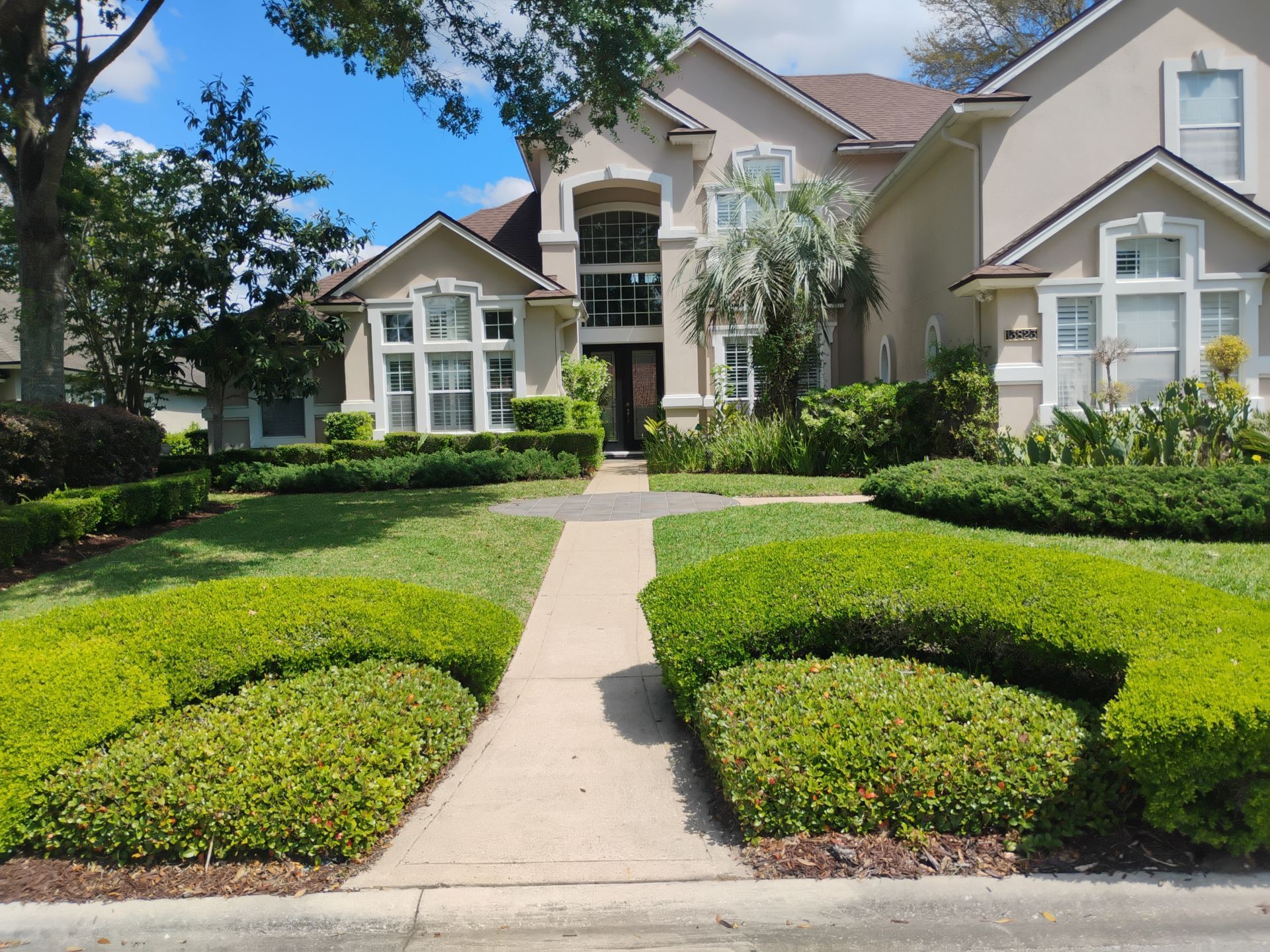 Large beige suburban house with a manicured front lawn, walkway, and trimmed hedges under a blue sky