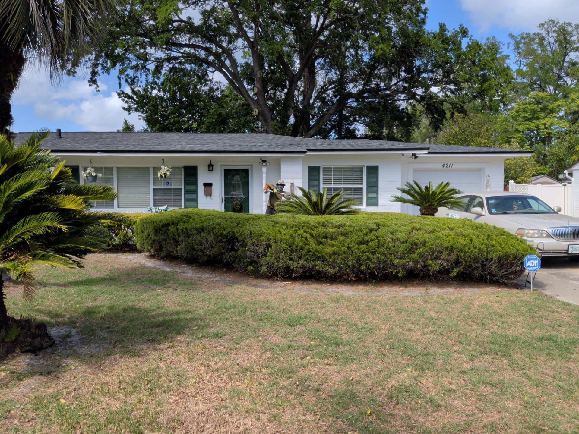 Single-story white house with front porch, trimmed hedges, and parked car under trees