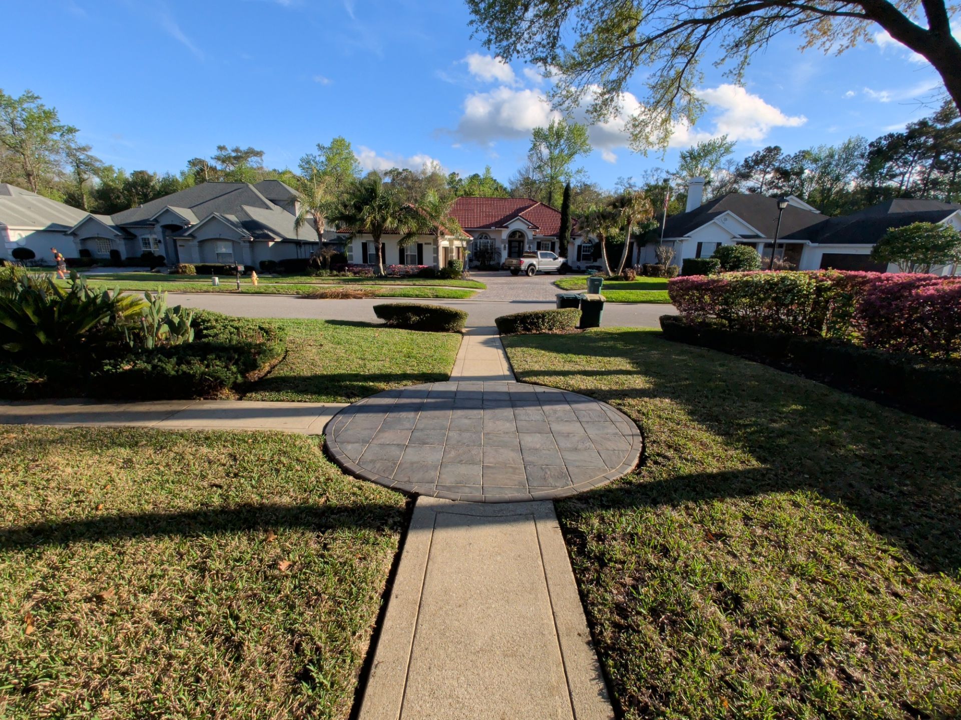 Suburban front walkway leading to a quiet street, with manicured lawns and houses under a blue sky.
