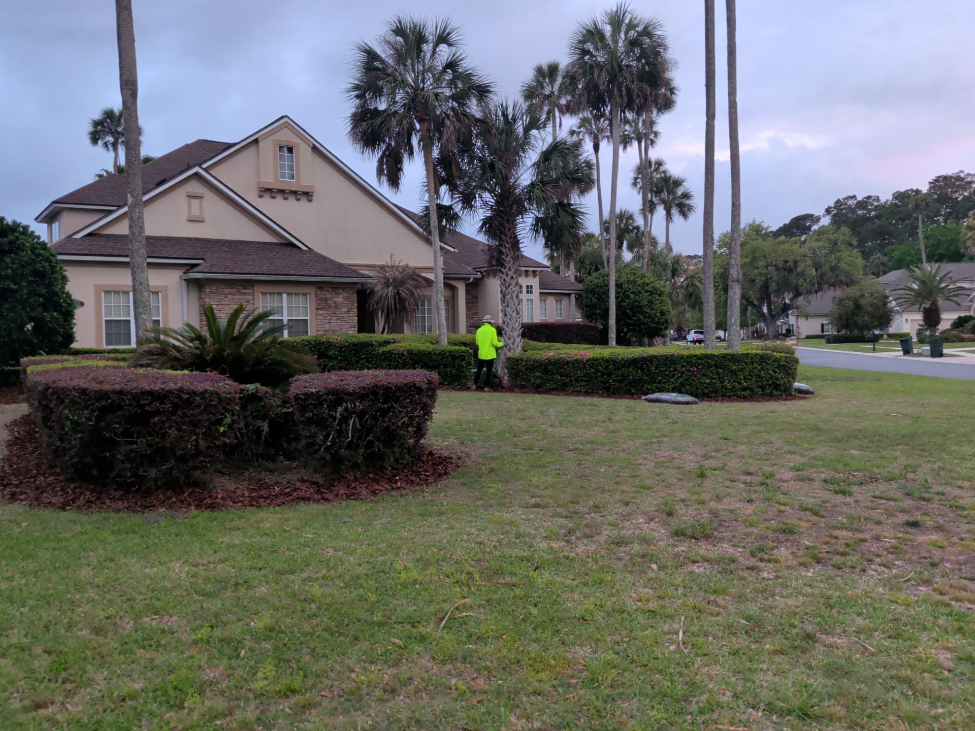 A suburban house with palm trees and a neon-clad worker standing on the lawn near a hedge.
