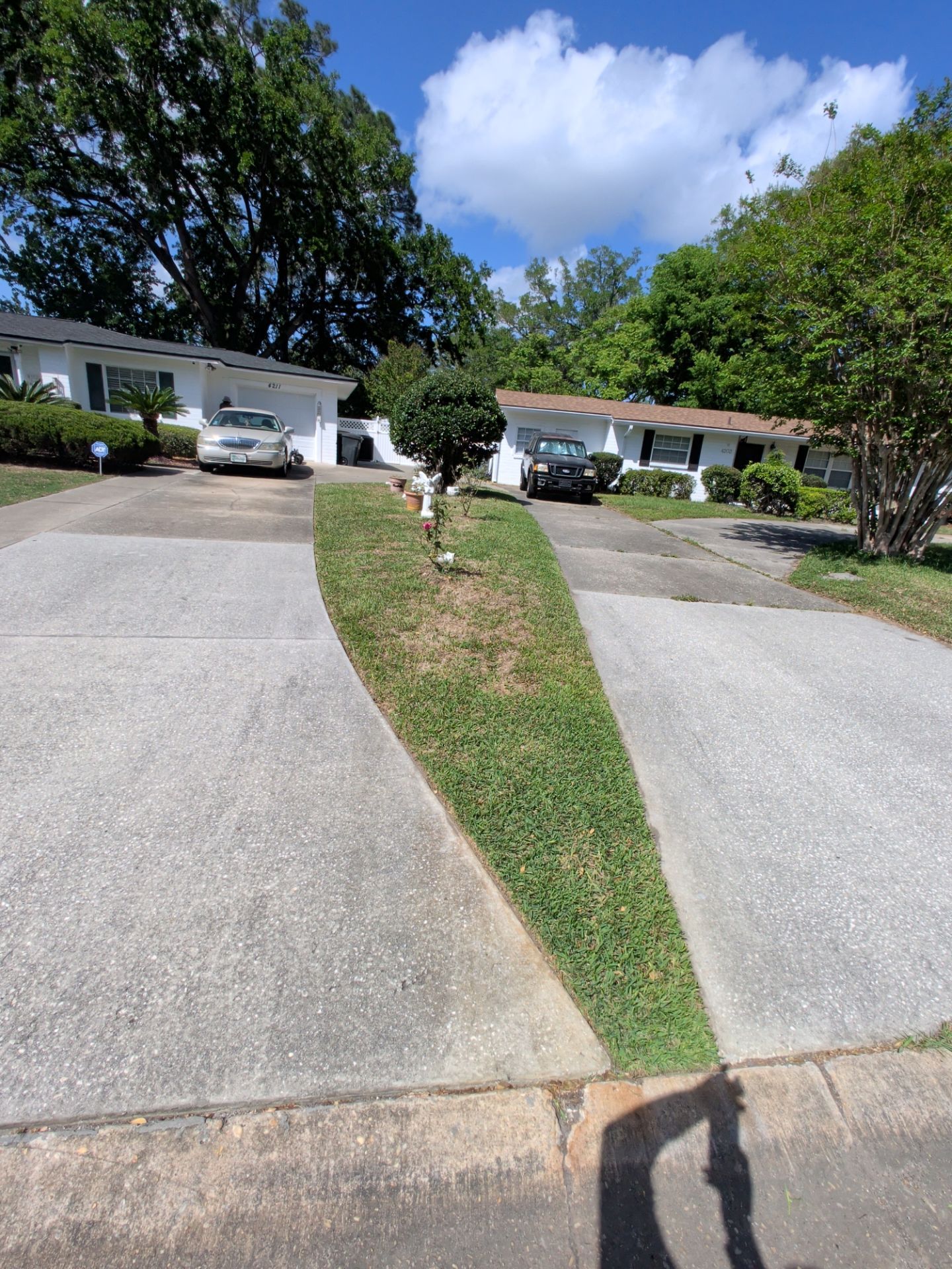 Suburban driveway split by a narrow grassy median, leading to white houses under a bright blue sky.