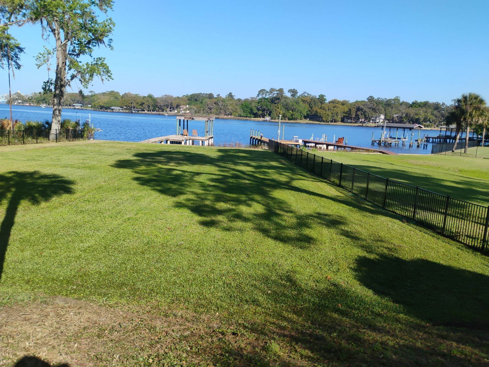 Sunny lakeside park with green lawn, shaded trees, and docked boats across the water