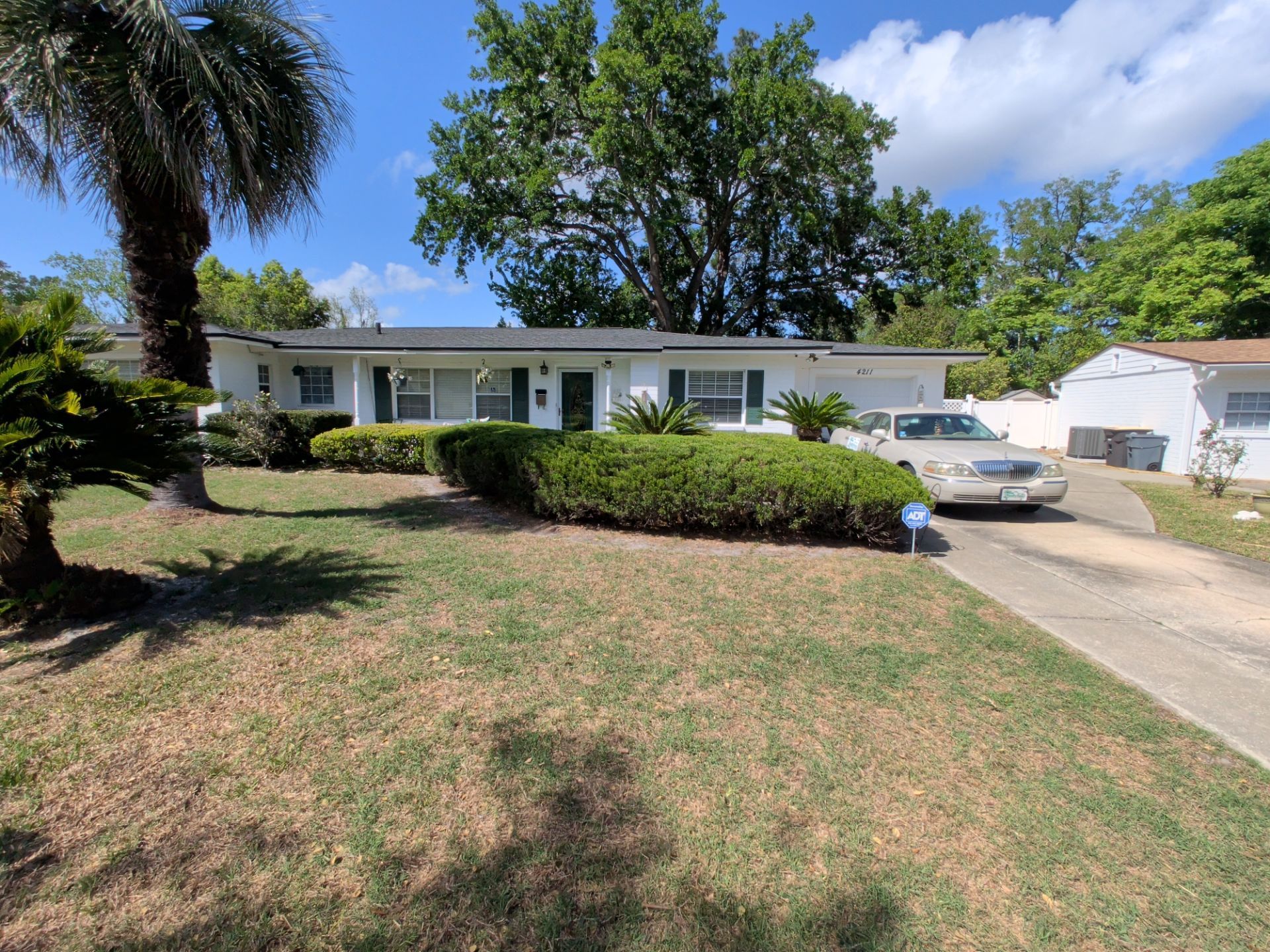 Single-story white house with a front lawn, shrubs, and a driveway with a parked car under a sunny sky